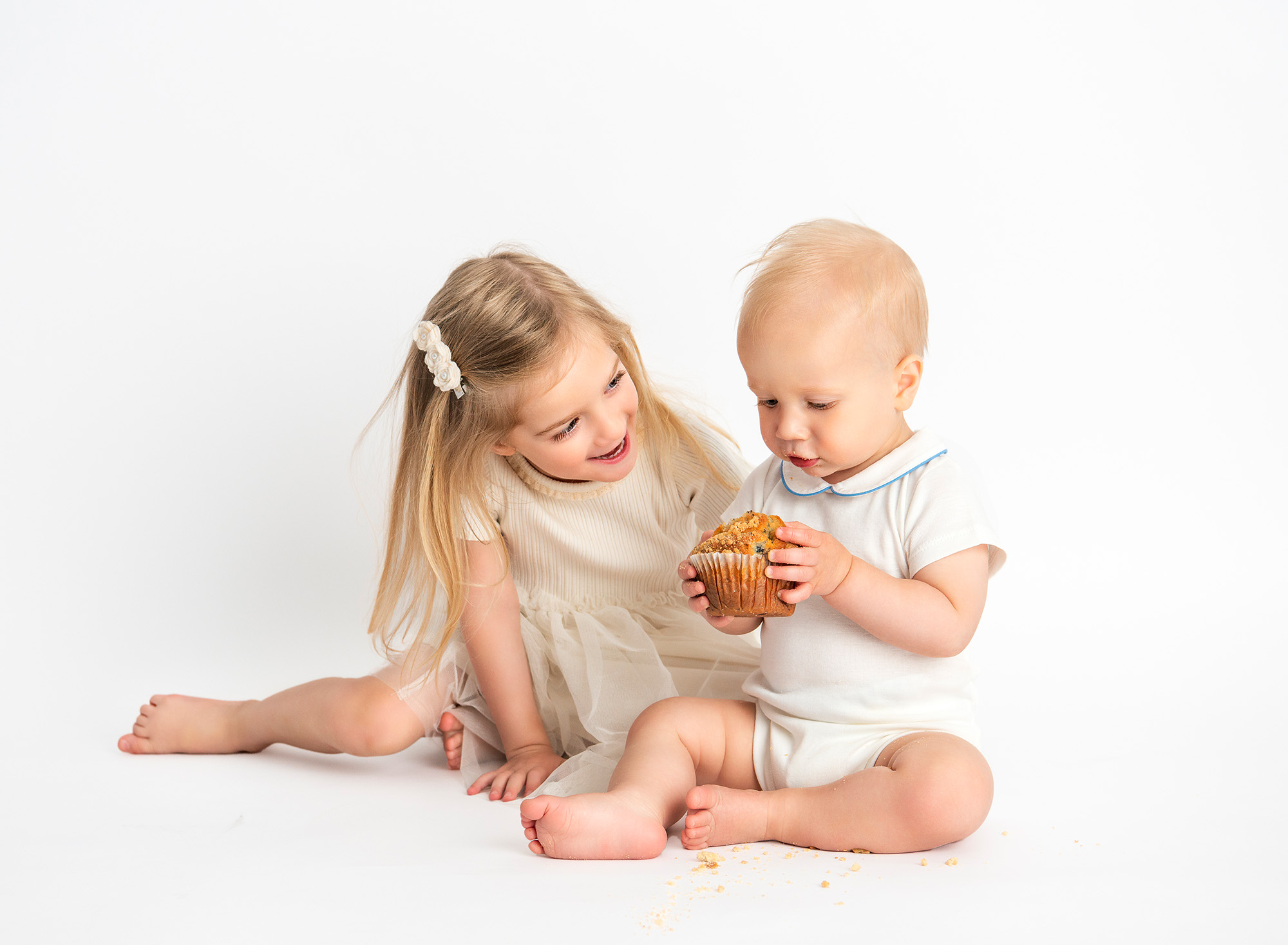 All White Baby Milestone Smash Photoshoot baby eating a blueberry muffin on a white background with his sister