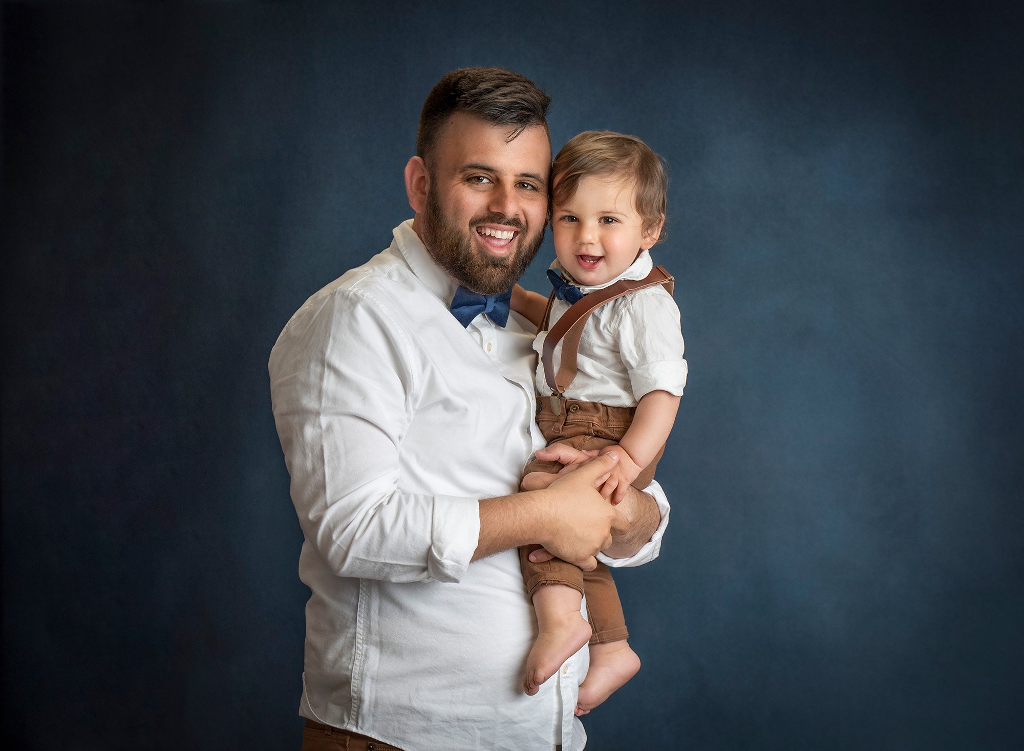 one year old boy in overalls smiling while being held by his dad on a blue background