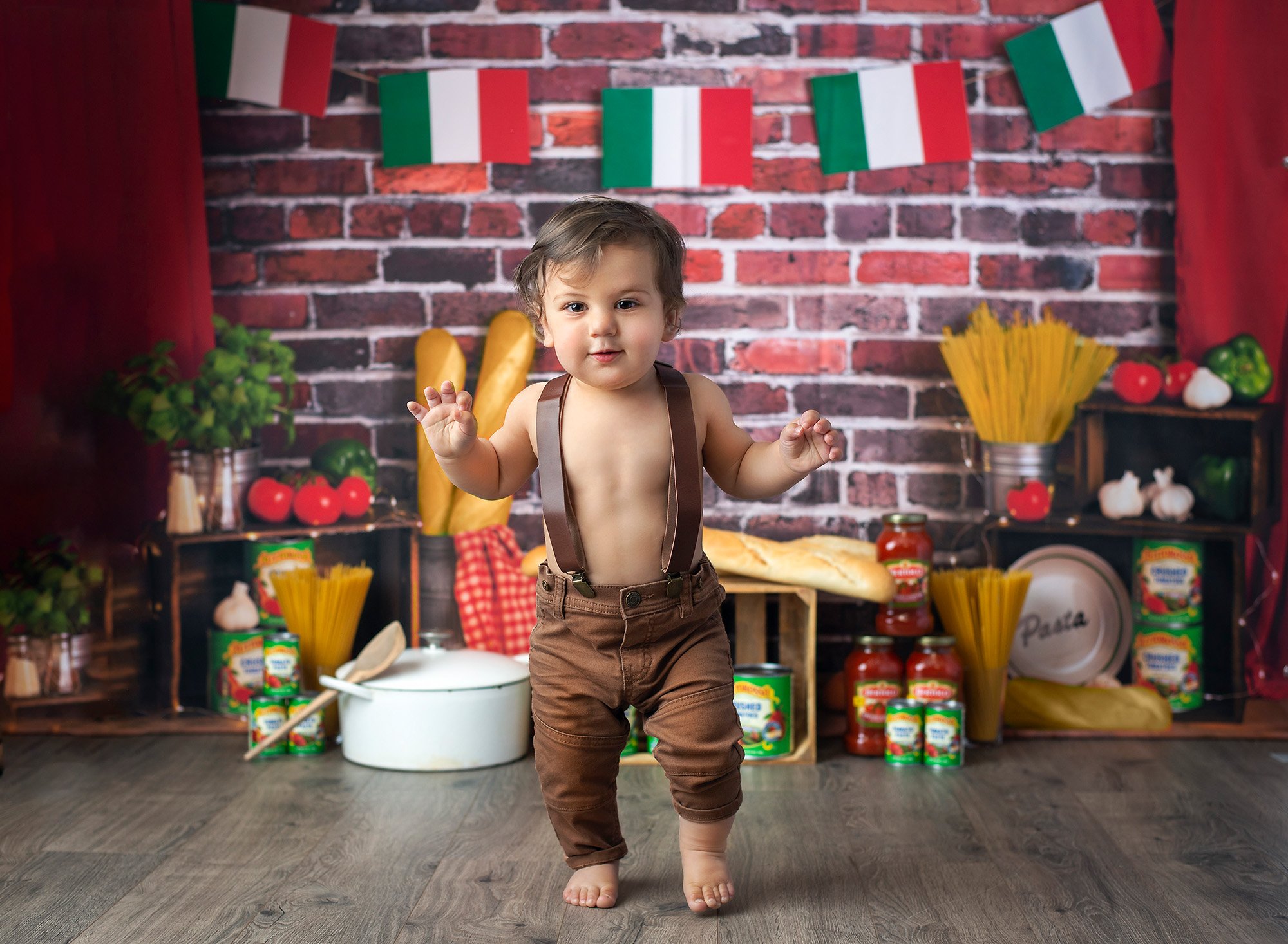 1 year baby photoshoot spaghetti smash one year old boy walking in overalls with Italian flags, Italian bread, pasta and jars of sauce in background