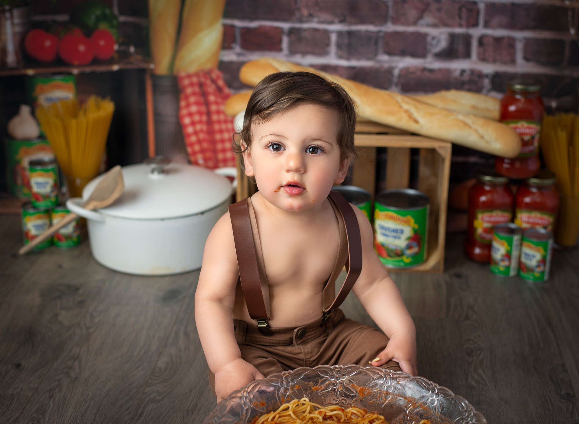 one year old boy wearing overalls with Italian bread and jars of sauce in the background