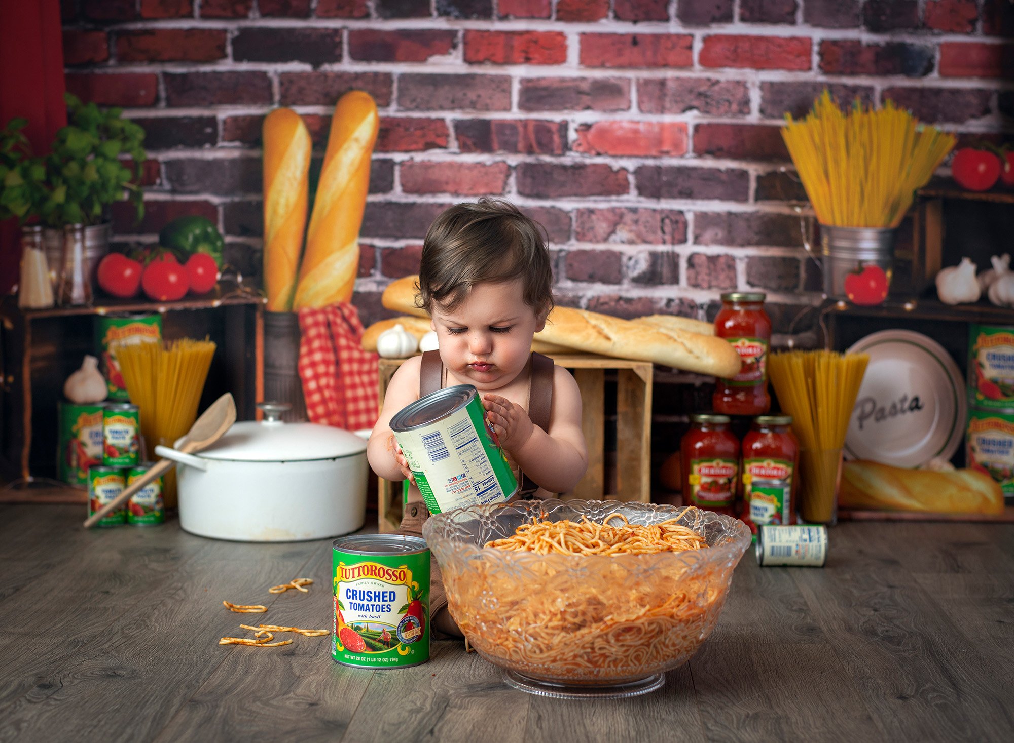 1 year baby photoshoot spaghetti smash one year old boy playing with tomato cans while surrounded by pasta and sauce jars on a brick background