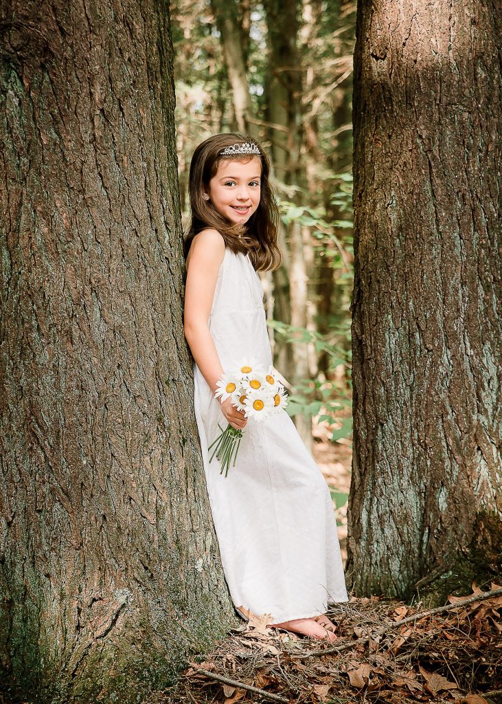 little brown haired girl holding white daisies in the forest leaning against a tree