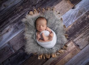 sleeping newborn boy swaddled in white lying in a rustic driftwood bowl on grey plank wood best time to take newborn photos