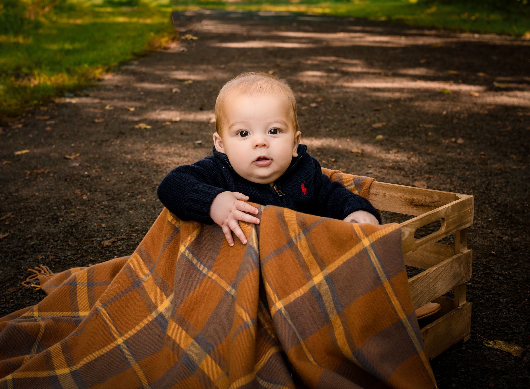 6 month old baby boy sitting in a crate with a fall blanket outside on a path