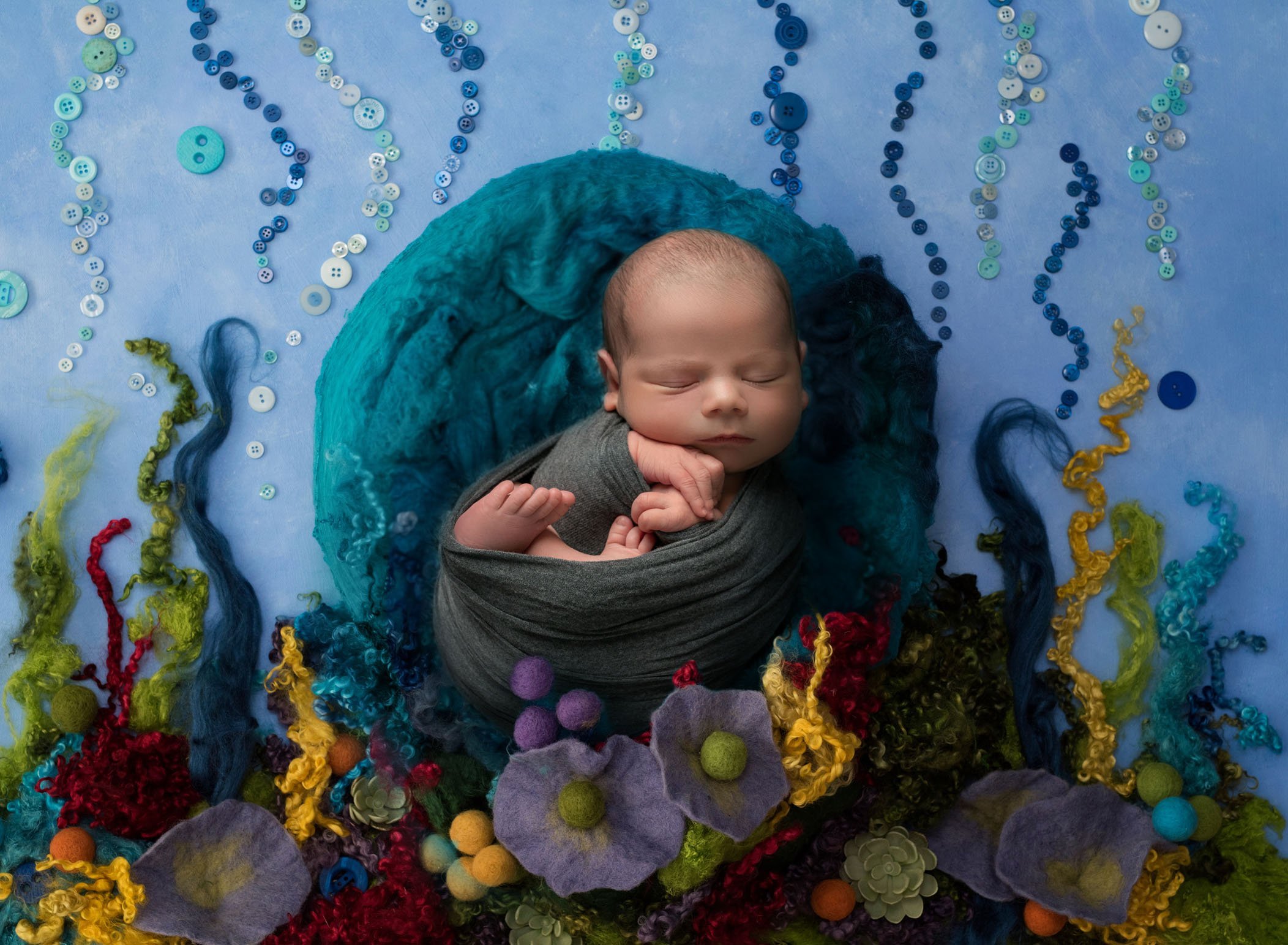 swaddled newborn sleeping in a blue wool nest among sea bead made out of felt and yarn