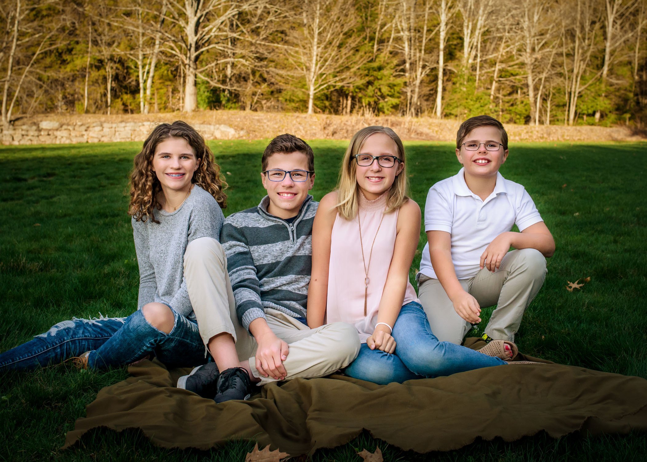 Four young siblings sit on grass in park smiling at camera