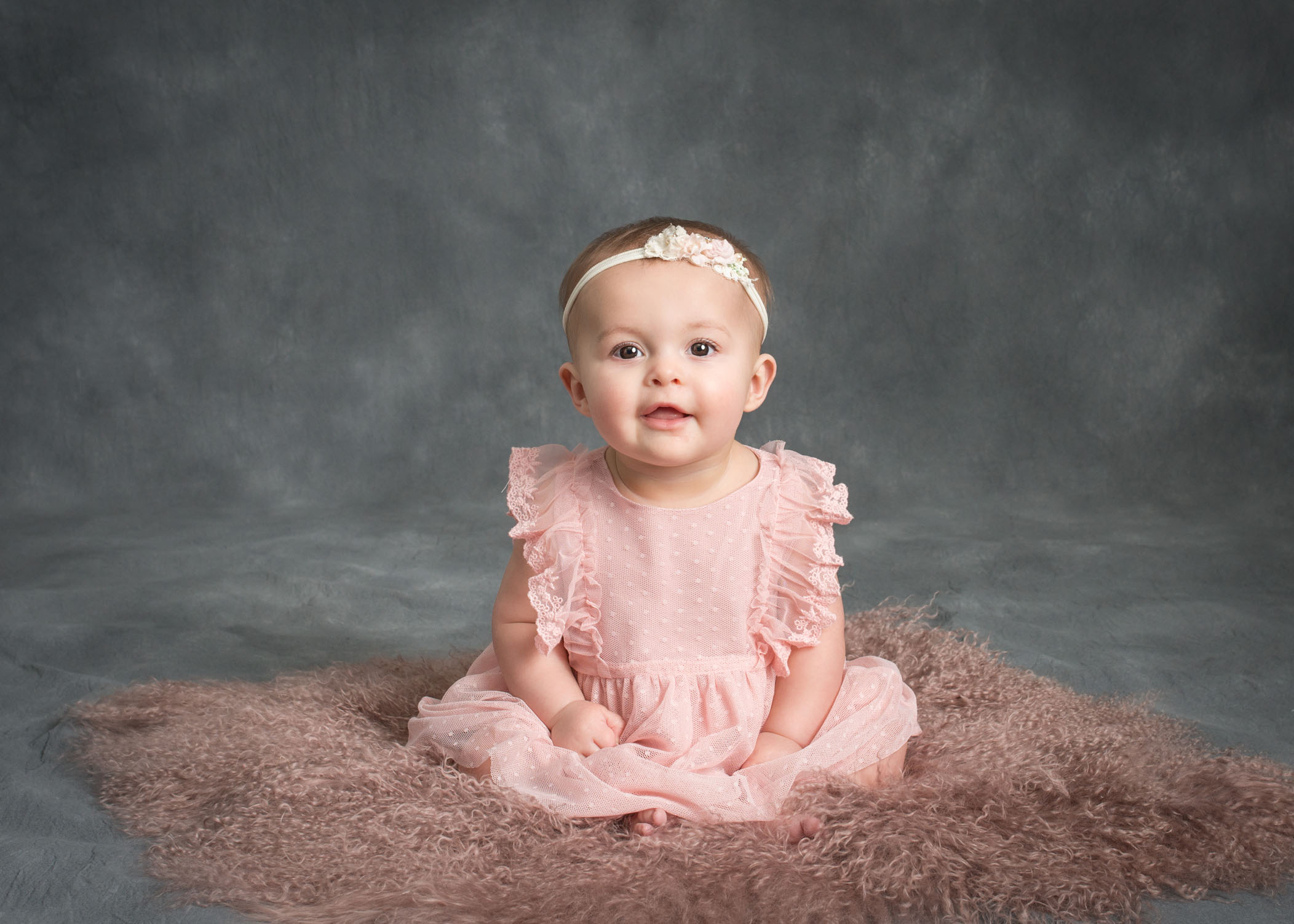6 month old baby girl smiling at camera sitting on pink fur on grey backdrop
