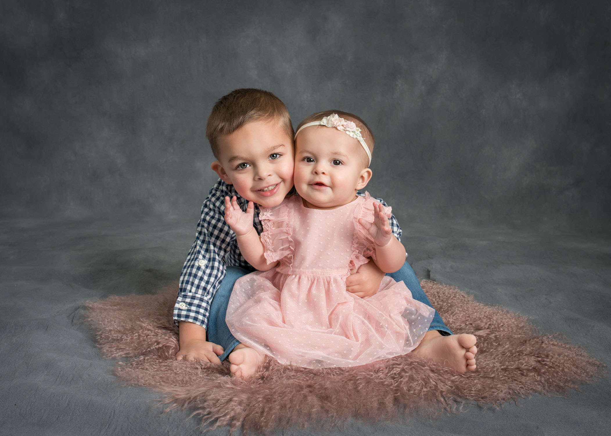 6 month old baby girl sitting on pink fur with 3 year old brother hugging her from behind and smiling