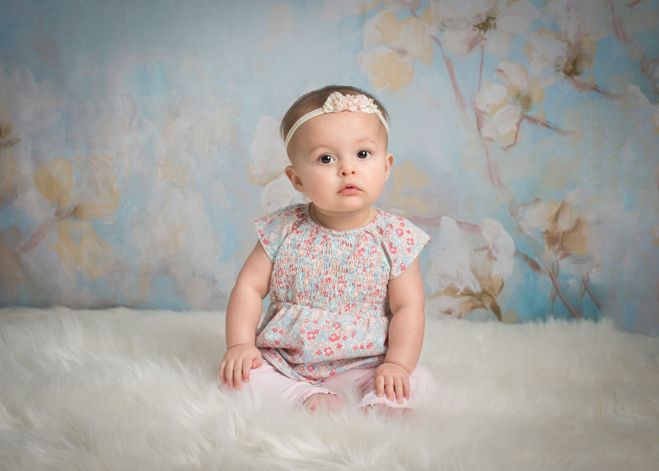 6 month old baby girl looking serious sitting on white fur in front of blue floral backdrop