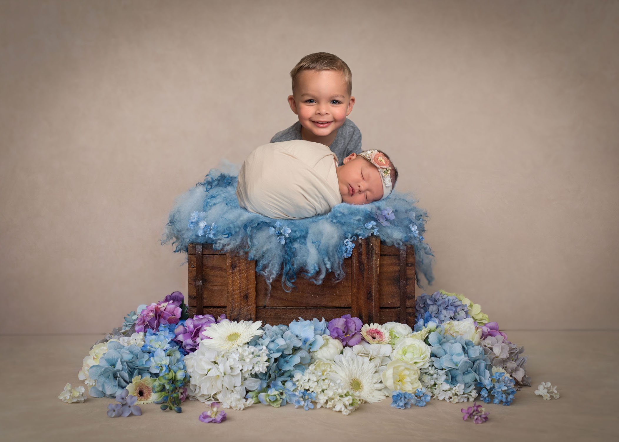 2 year old big brother proudly posing with newborn sister on a crate with flowers