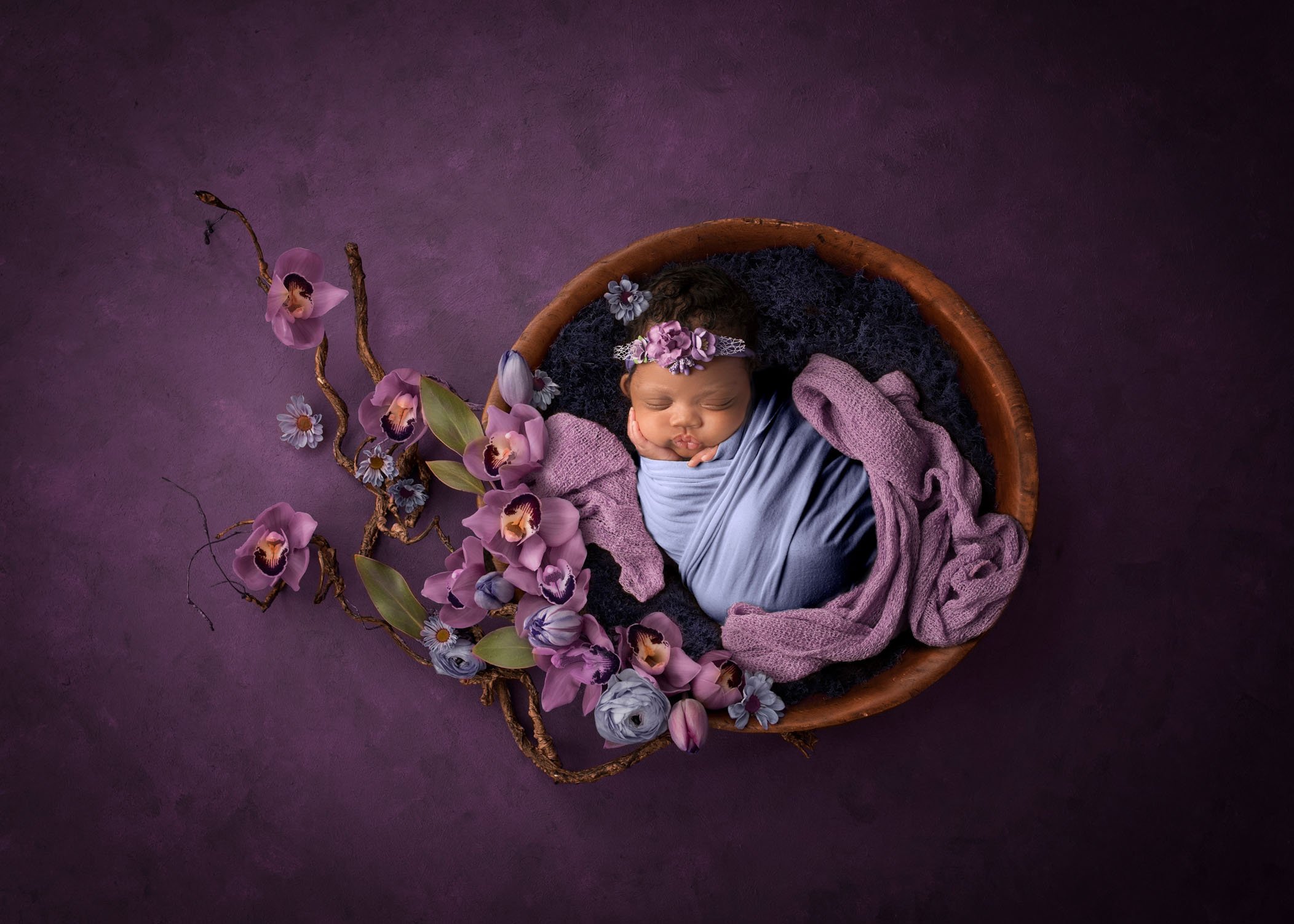 african-american newborn baby girl asleep in a bowl on a purple backdrop with purple orchids surrounding her