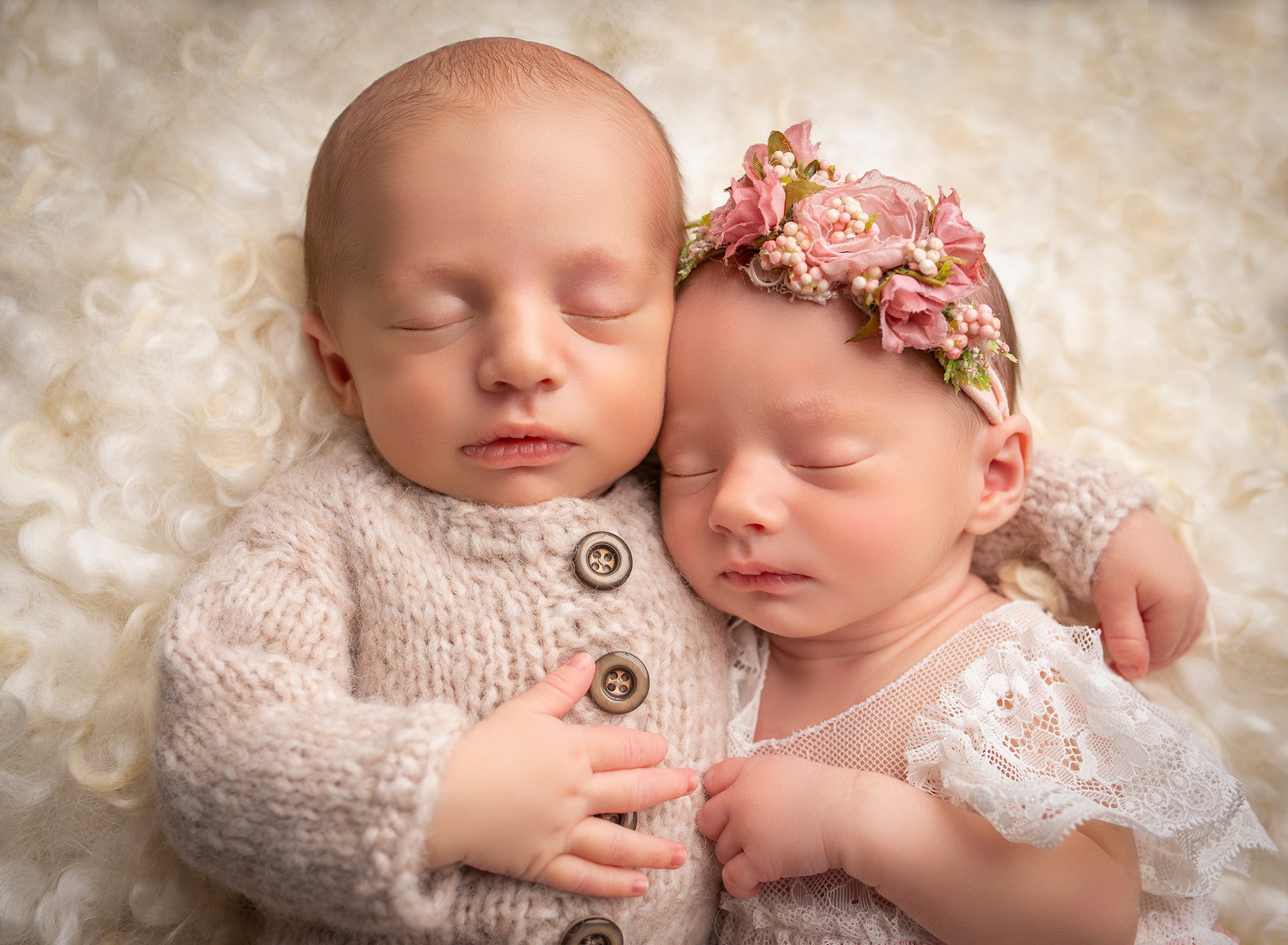 Twins newborn photography newborn baby boy asleep with his arm wrapped around his twin sister asleep in lace dress with pink floral headband on white fuzzy blanket