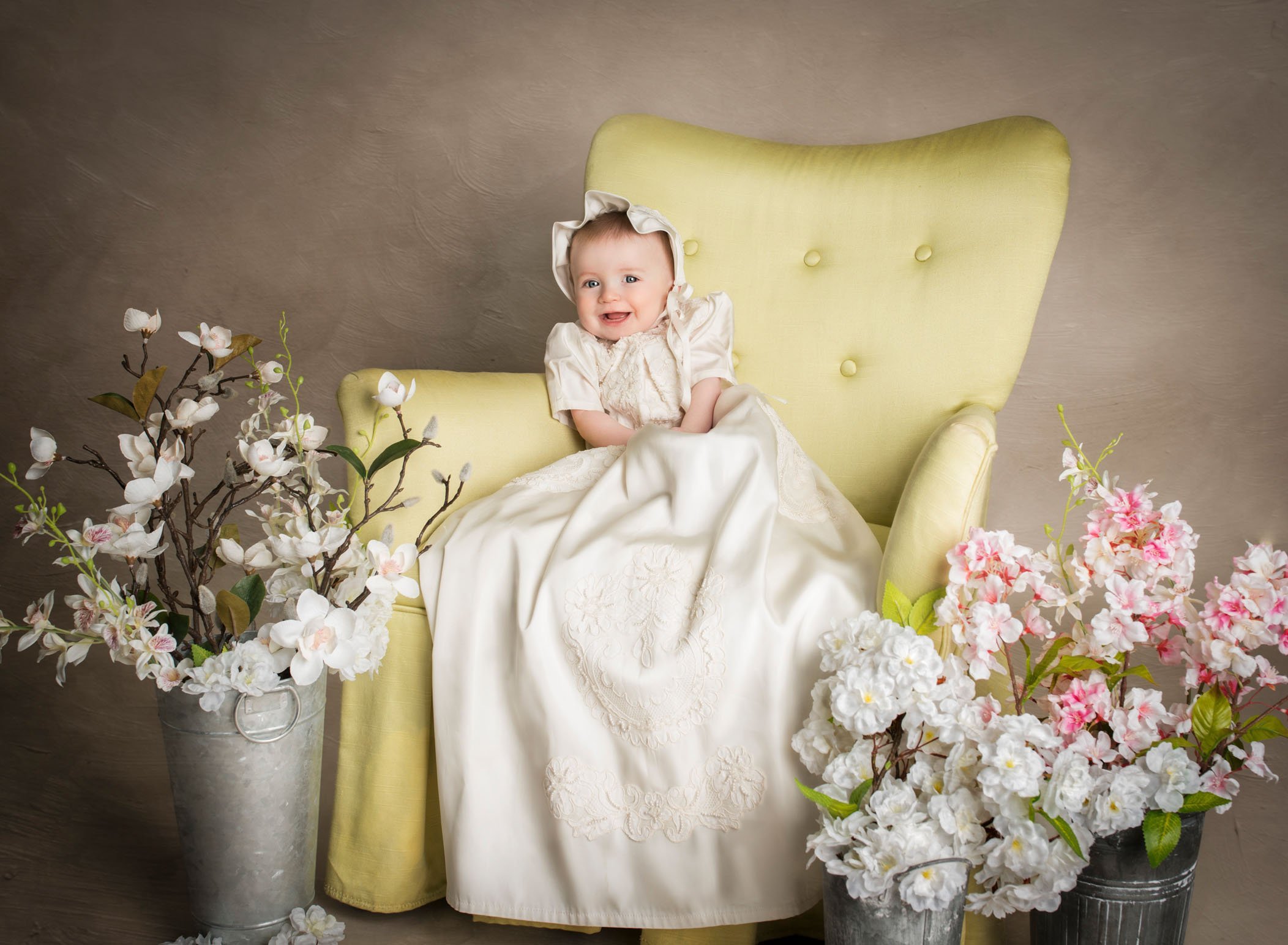 6 mo old baby girl sitting in a chair surrounded by flowers dressed in her fancy Christening gown