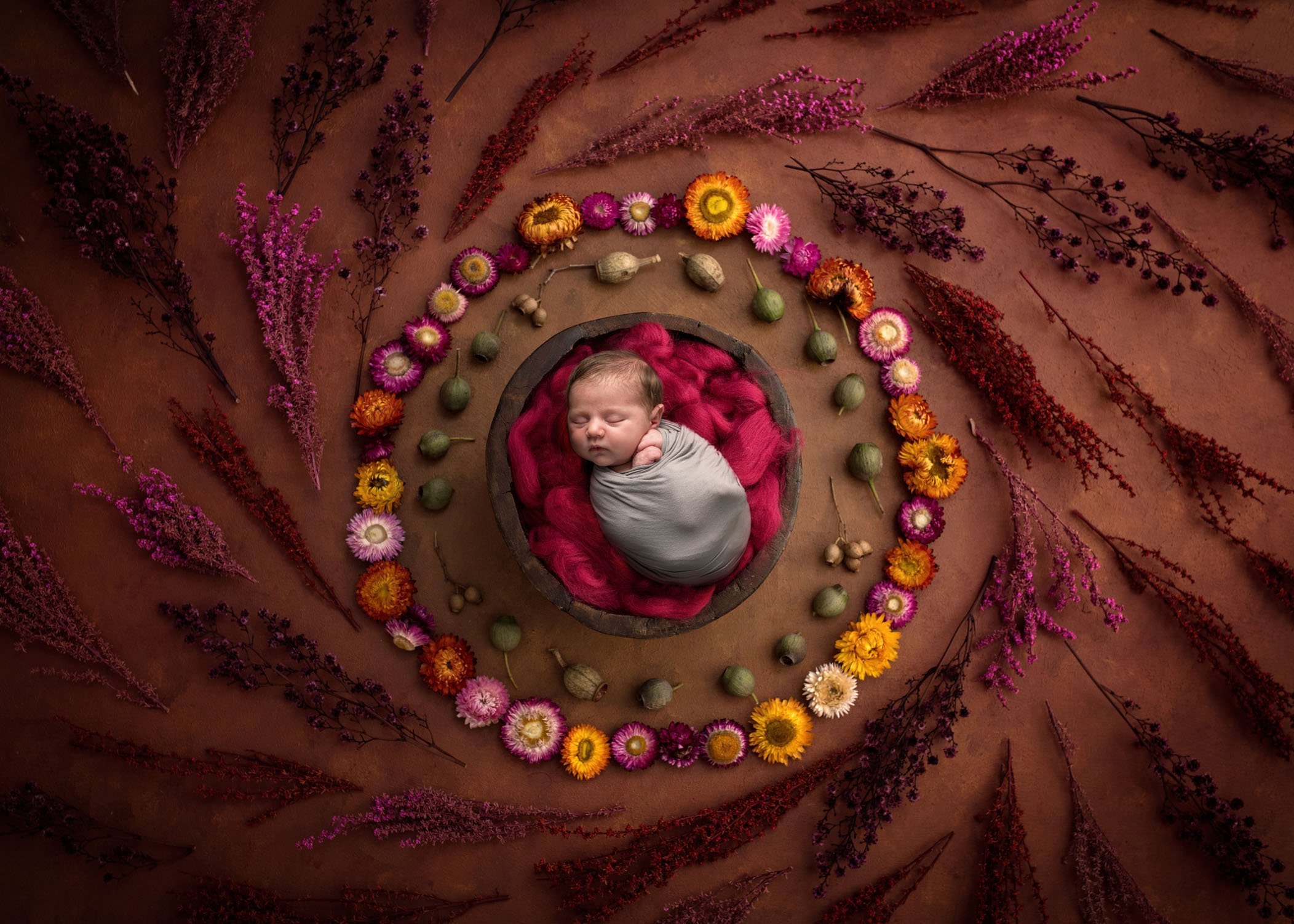 newborn baby sleeping in red bowl of fluff surrounded by swirl of flowers
