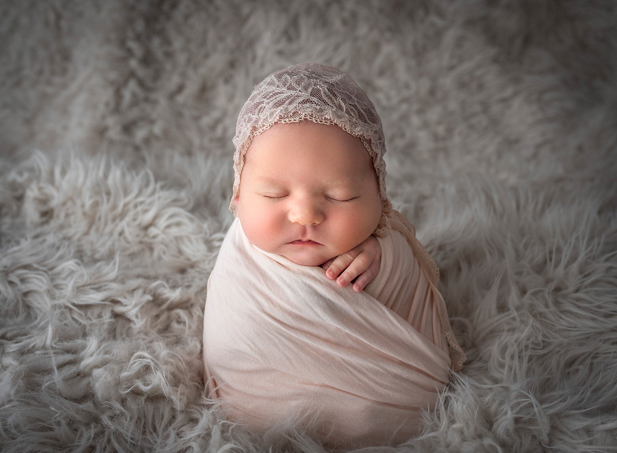 newborn baby girl sound asleep in soft pink wrap and laced bonnet on a fluffy blanket