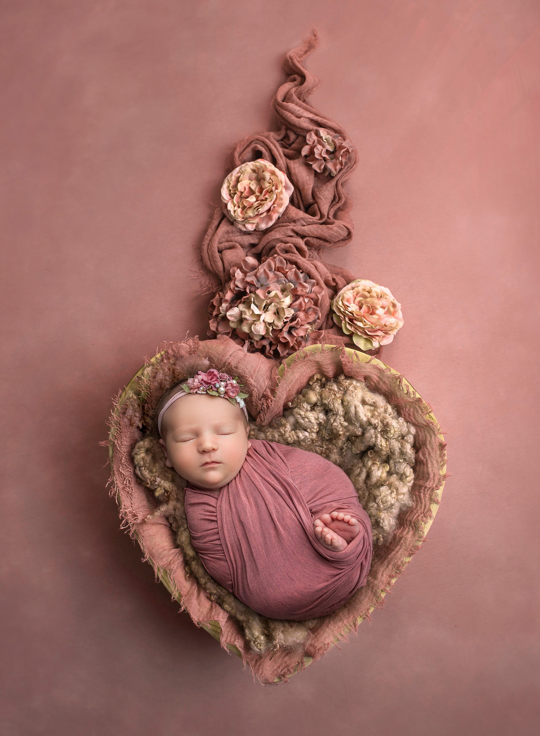 newborn baby girl asleep in mauve wrap laying inside heart basket with flowers