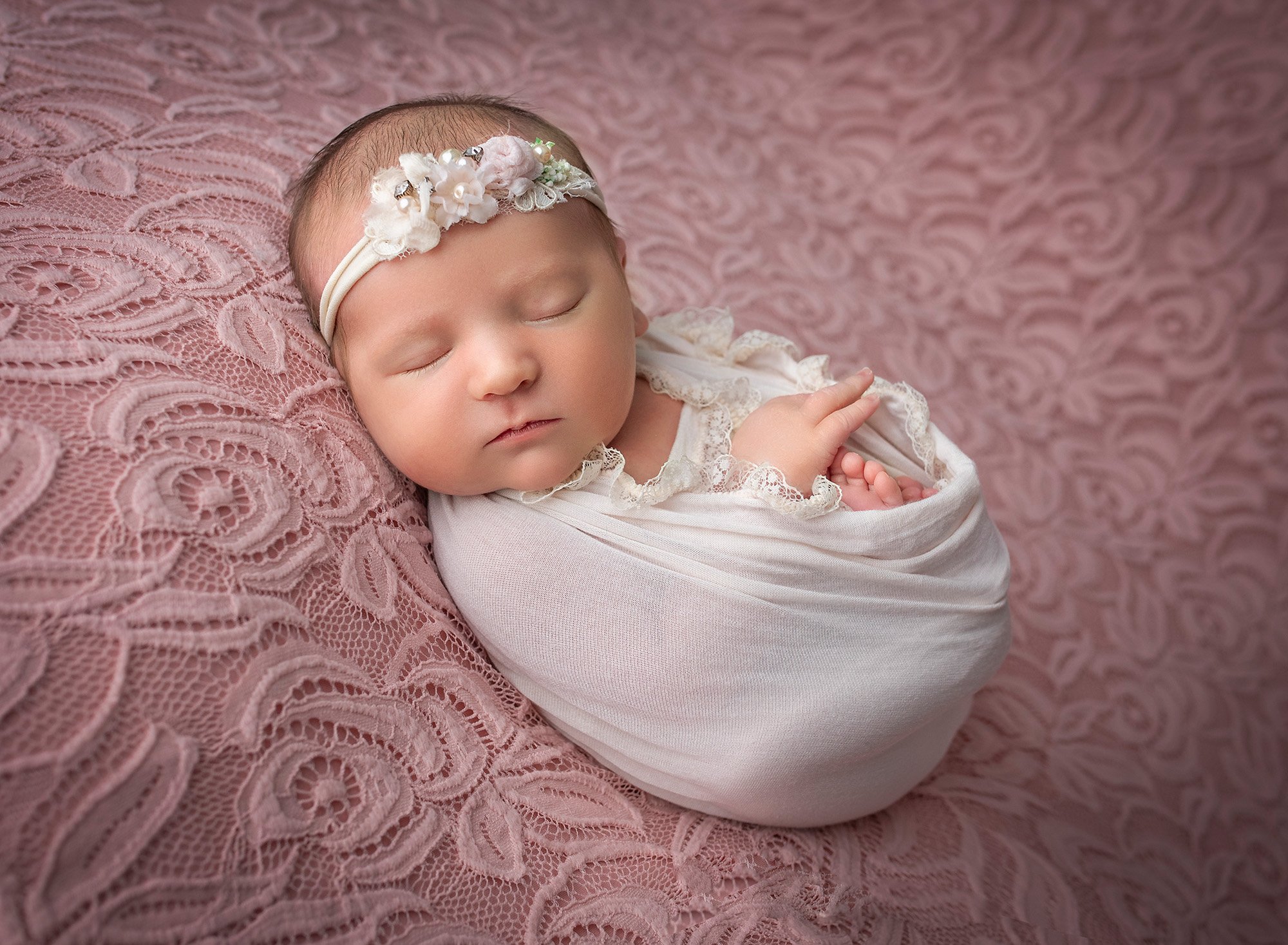 newborn baby girl swaddled in lace white wrap asleep on laced pink background