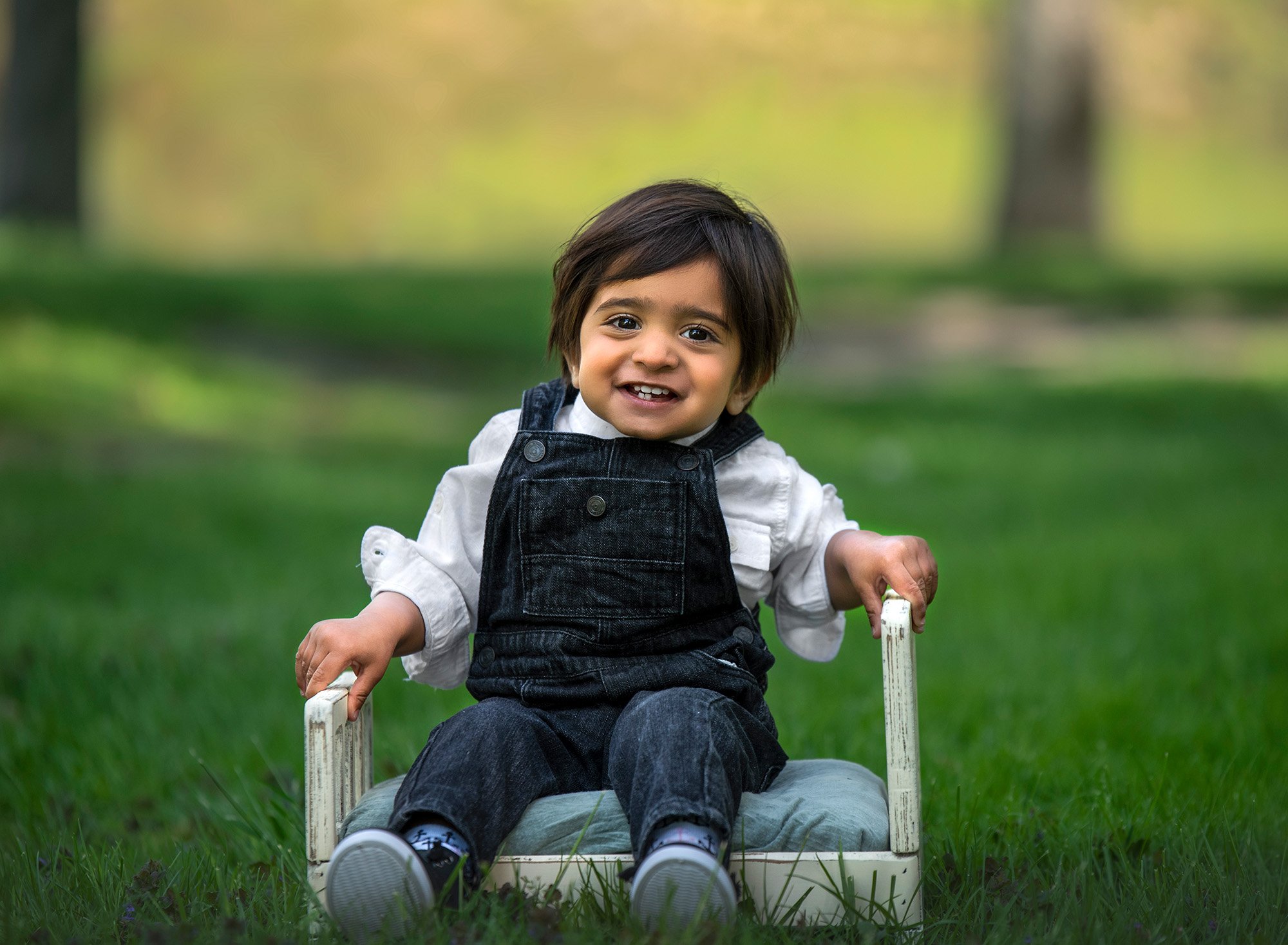 Springtime One Year Photos little boy dressed in overalls sitting on miniature white bed with blue cushion