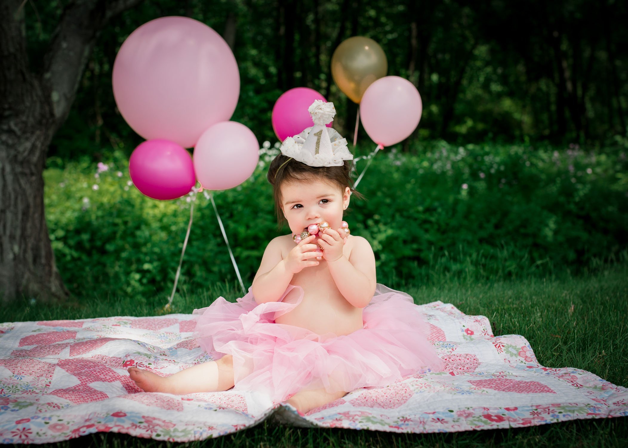 one year old girl chewing on a necklace wearing a tutu with balloons in the background