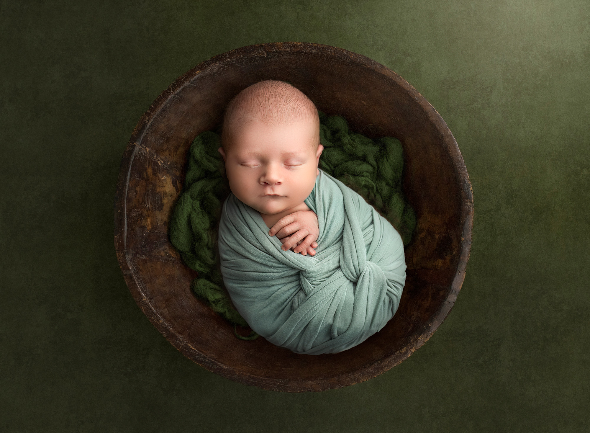 Owen asleep in a wooden bowl on a braided blanket, wrapped in green