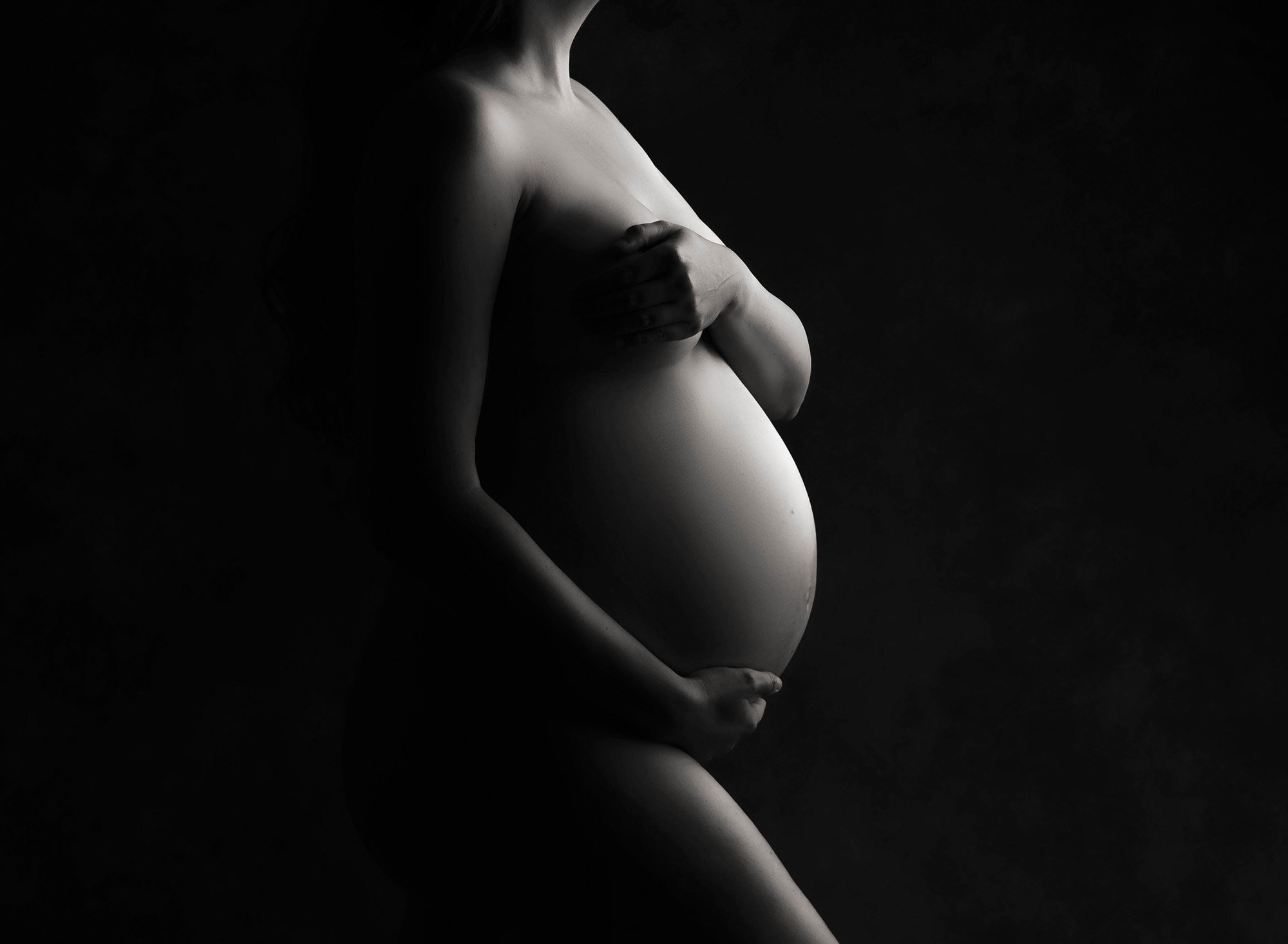 black and white photo of naked pregnant woman from her neck down, holding her stomach and cupping her breast