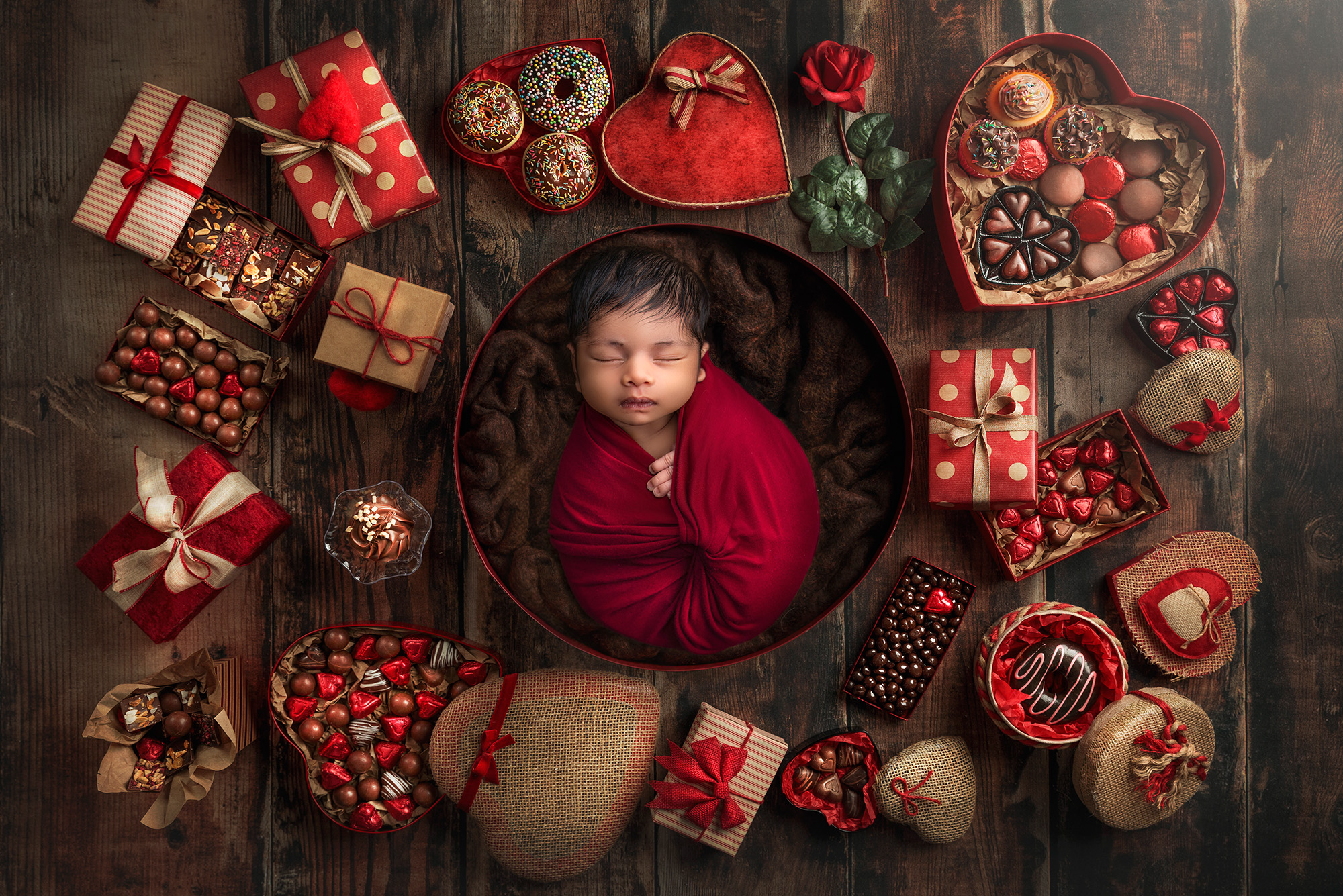 newborn baby boy wrapped in a cranberry red sleeping in a round box surrounded by red and gold wrapped gifts and candies Fine Art Newborn Photography