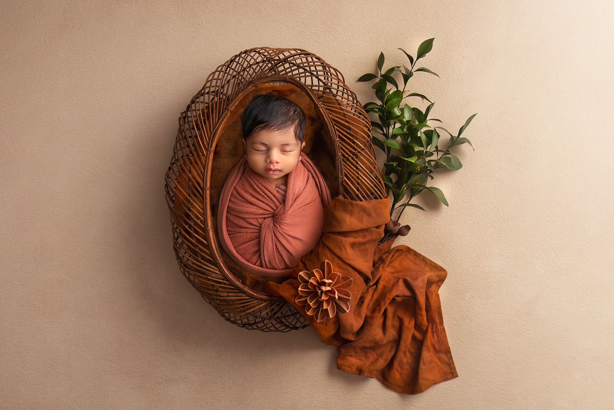 baby boy wrapped in a rust colored fabric sleeping in a wicker bowl with a green sprig and a flowing wrap next to him Fine Art Newborn Photography