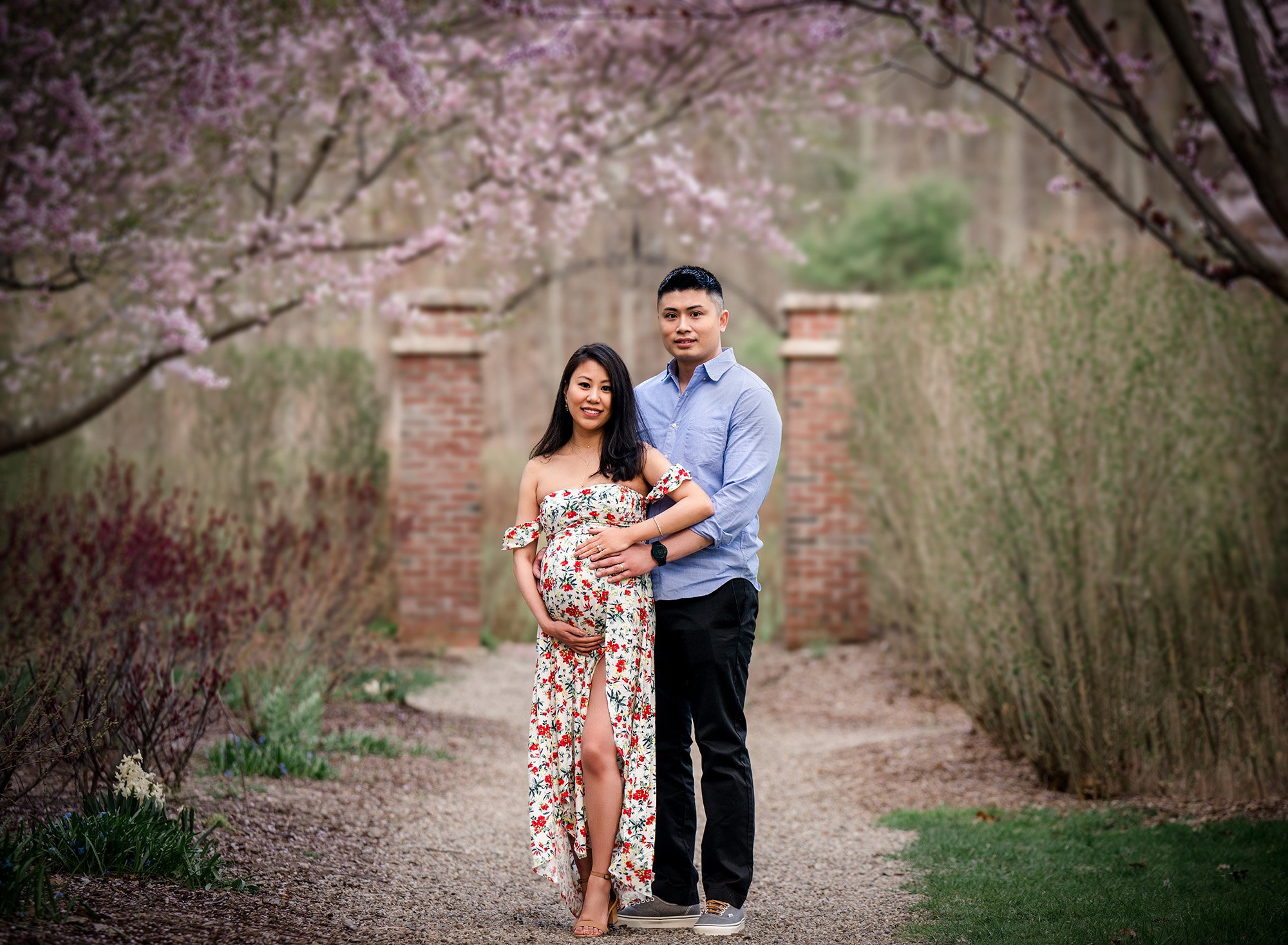 pregnant woman in floral maternity dress posing with partner under weeping cherry trees in front of a brick archway