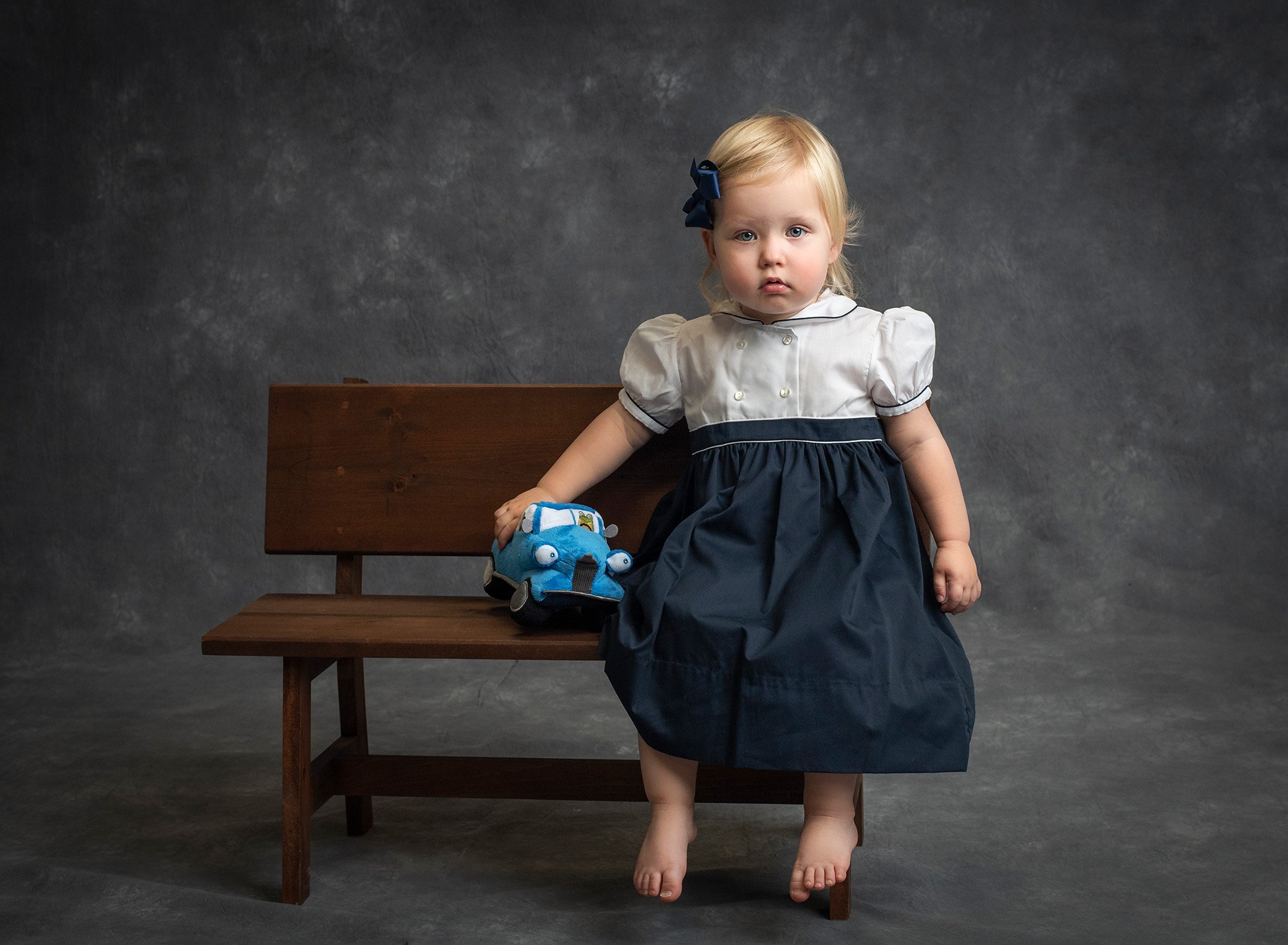 little girl sitting on wooden bench wearing a vintage dress holding a stuffed animal car