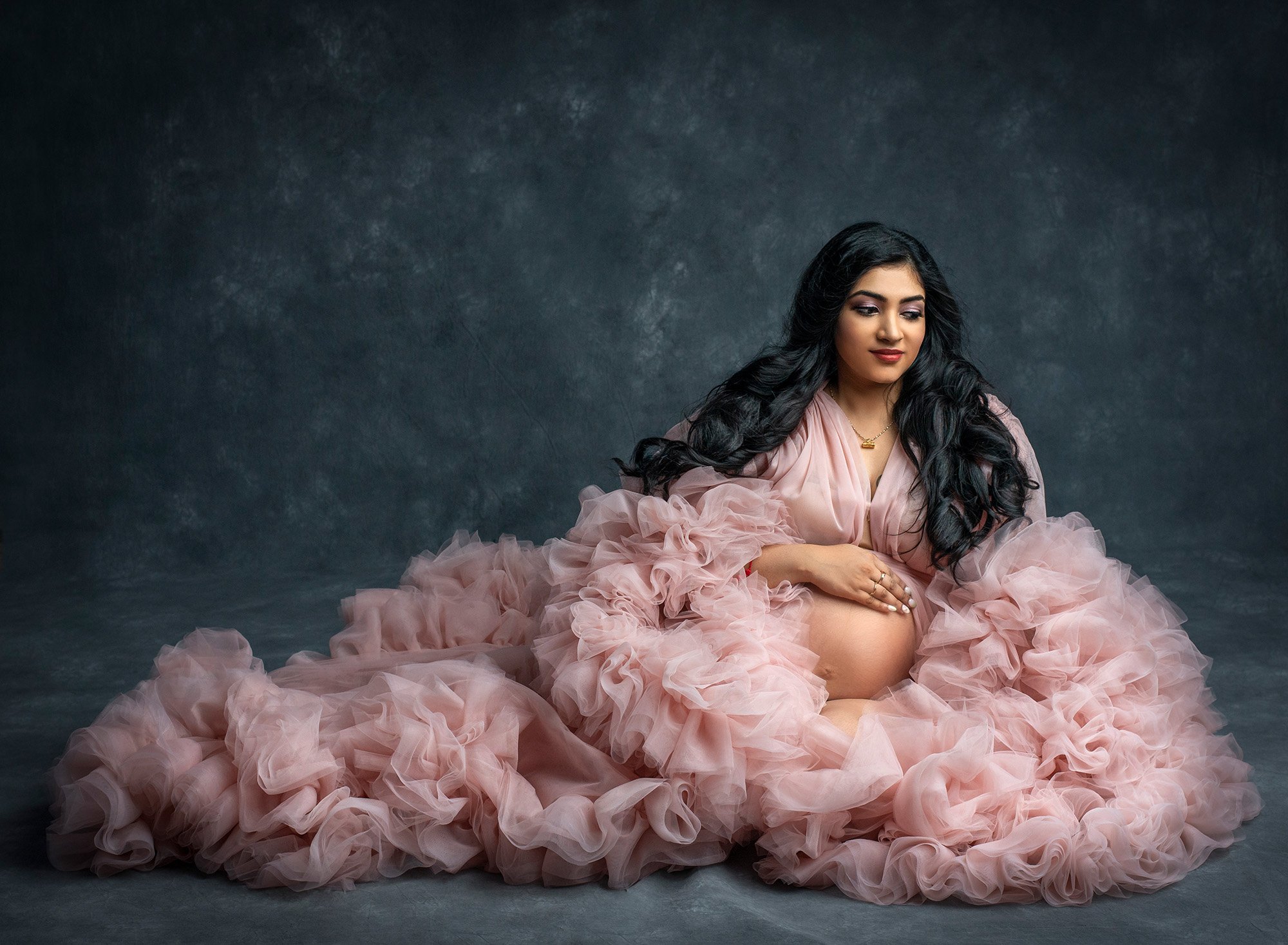 Indian Goddess Maternity photographs pregnant woman sitting down wearing ruffled pink dress exposing pregnant stomach