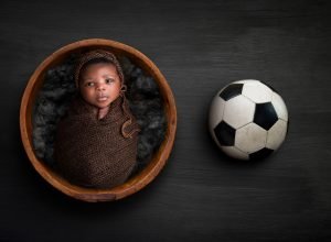 newborn baby boy wide awake swaddled in brown inside of wooden bowl next to a soccer ball