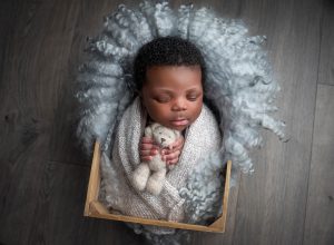 Jamaican boy newborn photographs newborn baby boy asleep on fuzzy blanket holding teddy bear inside of a wooden crate