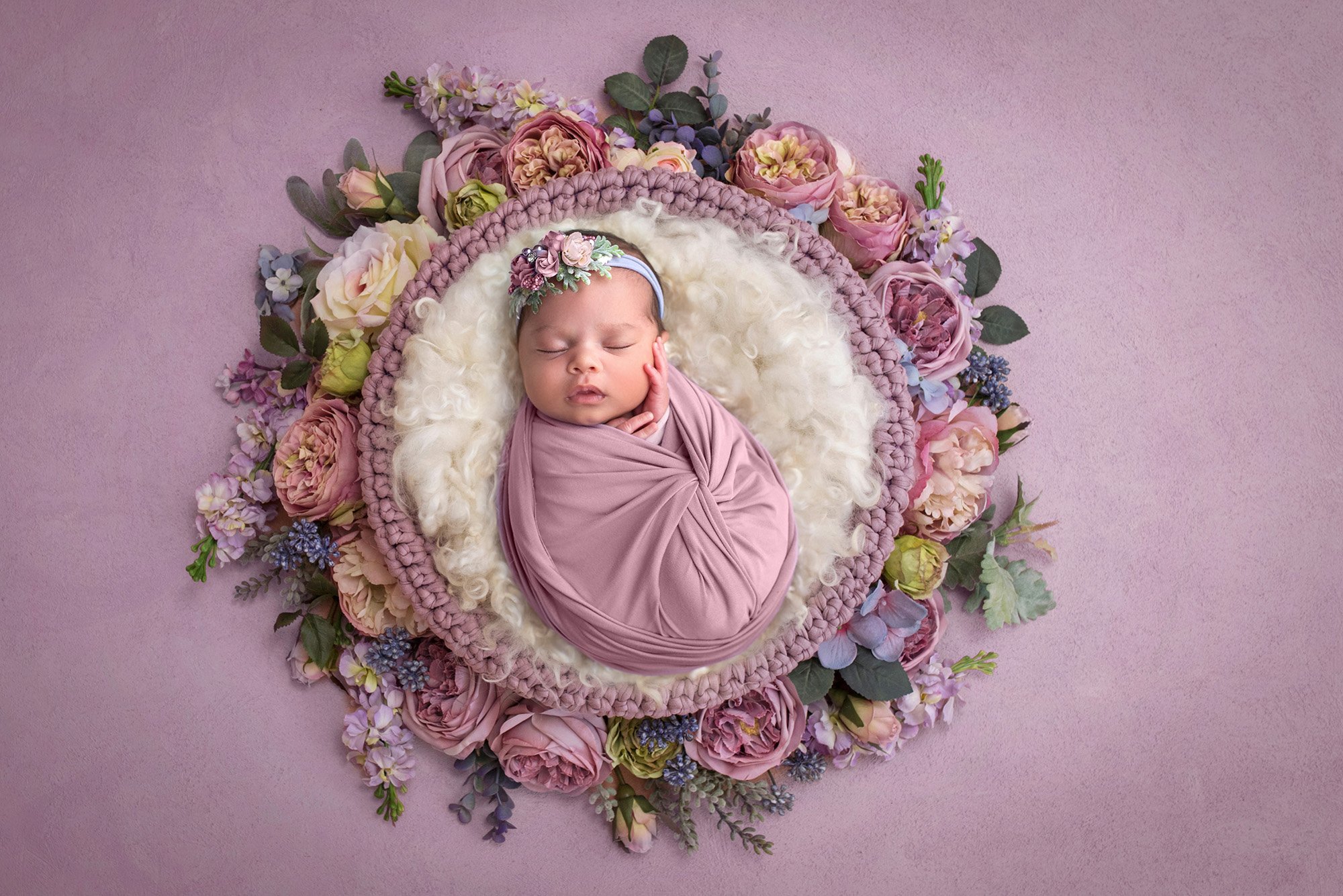 newborn baby girl swaddled in purple laying on white fuzzy blanket inside a wreath of flowers on purple backdrop