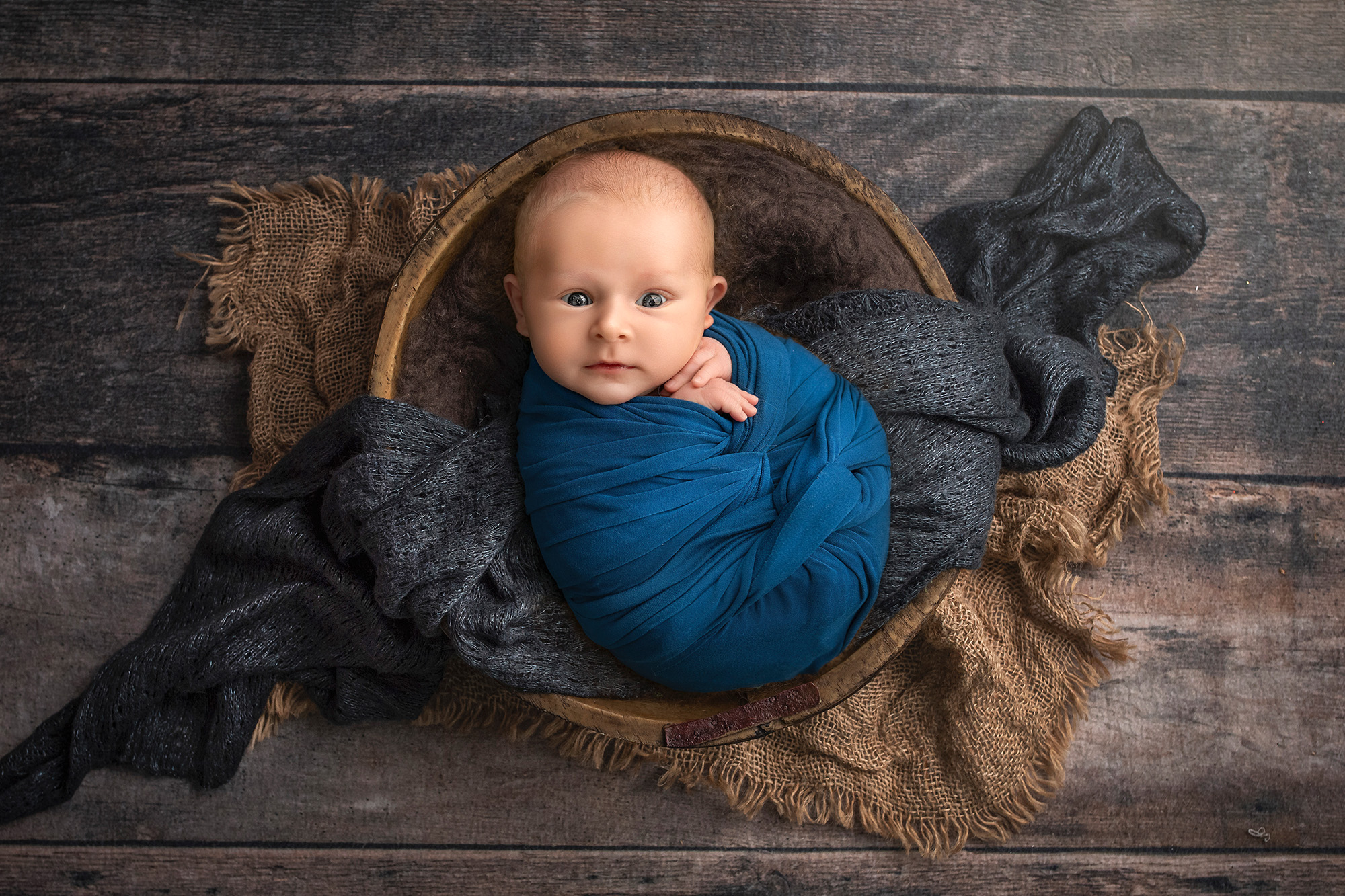 Awake newborn Levi swaddled in teal, nestled in a wooden bowl with layered fabrics.