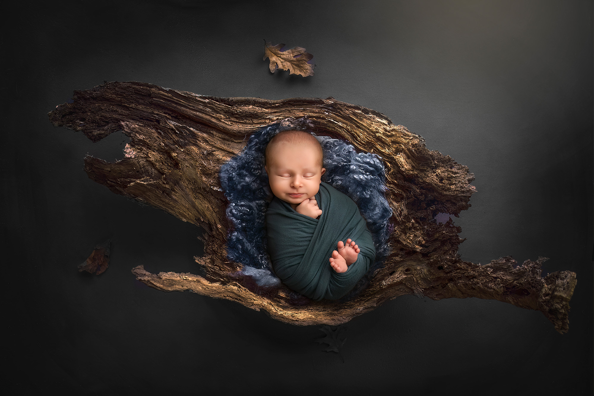 Sleeping newborn Levi in deep green wrap, resting in a natural wood prop with curly wool.