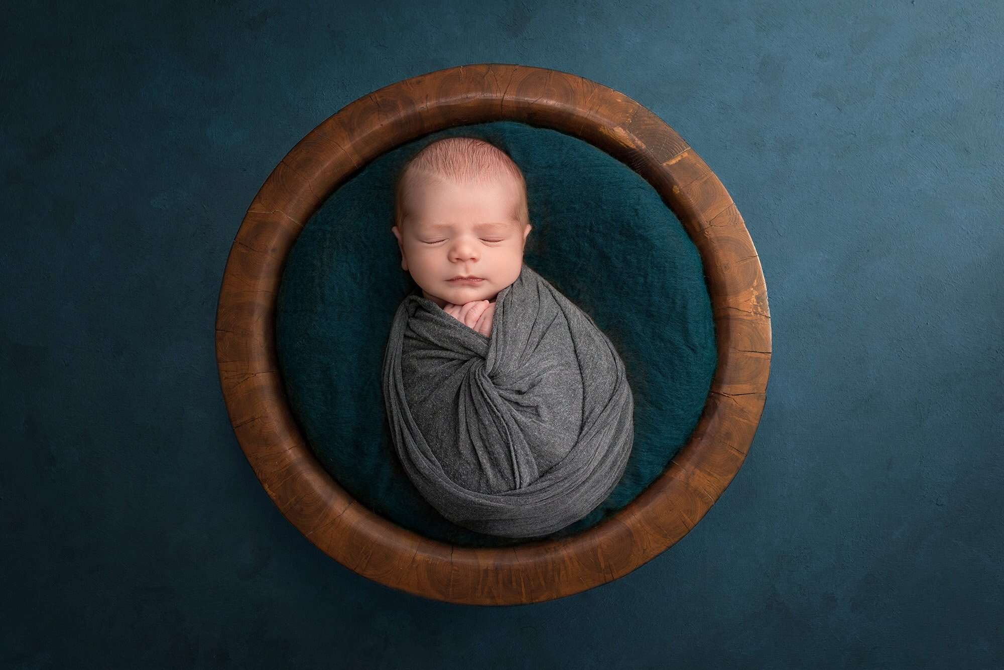 newborn baby boy swaddled in gray laying in wooden bowl with teal background