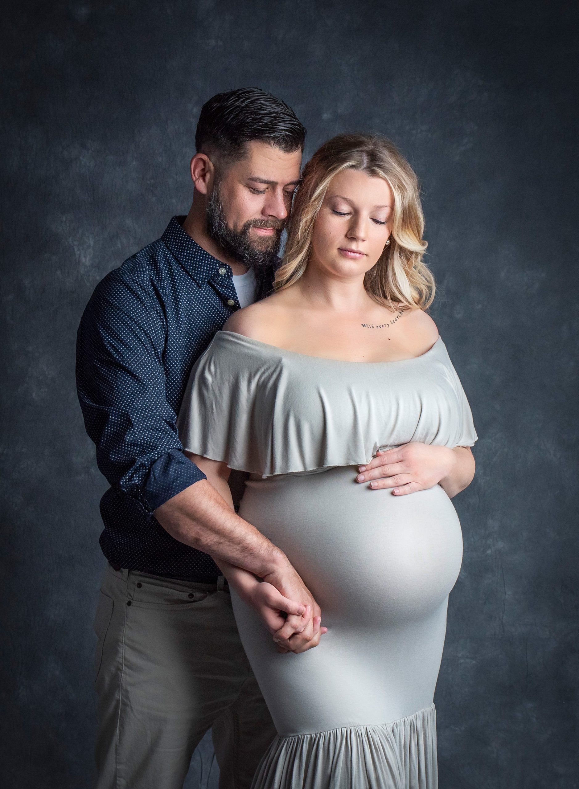 blonde pregnant woman posing in grey maternity dress while holding hands with her partner