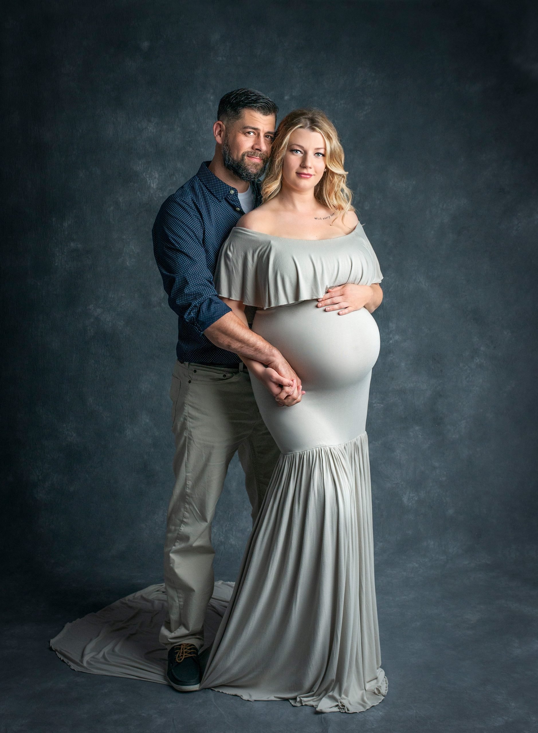 blonde pregnant woman posing in grey maternity dress while holding hands with her partner