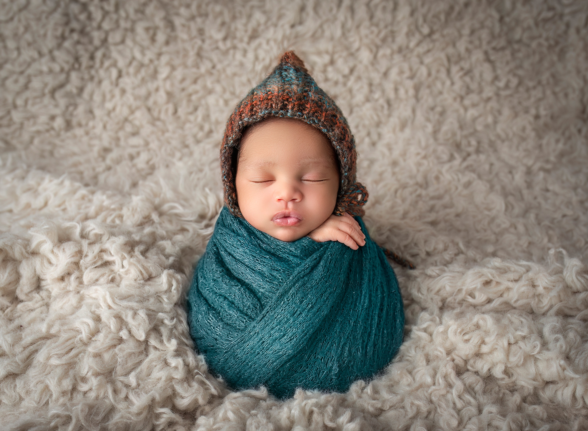 Newborn boy wrapped in teal wearing a multi colored bonnet sleeping on an oatmeal colored fur