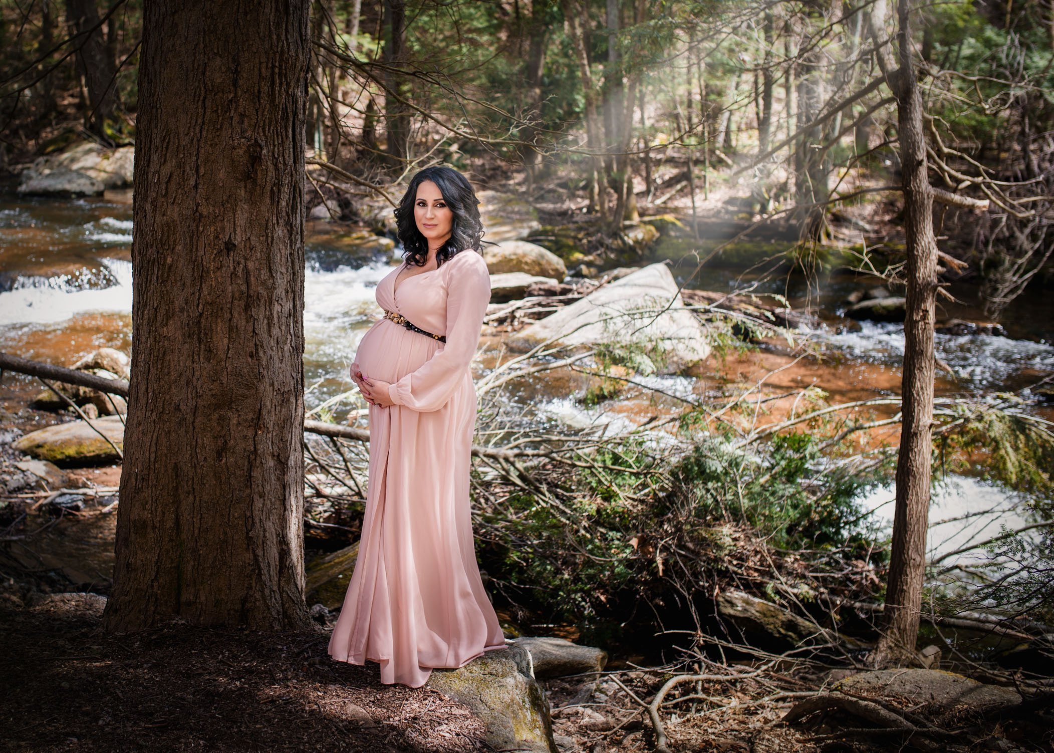 pregnant woman poses in the woods in front of a river with sunbeams