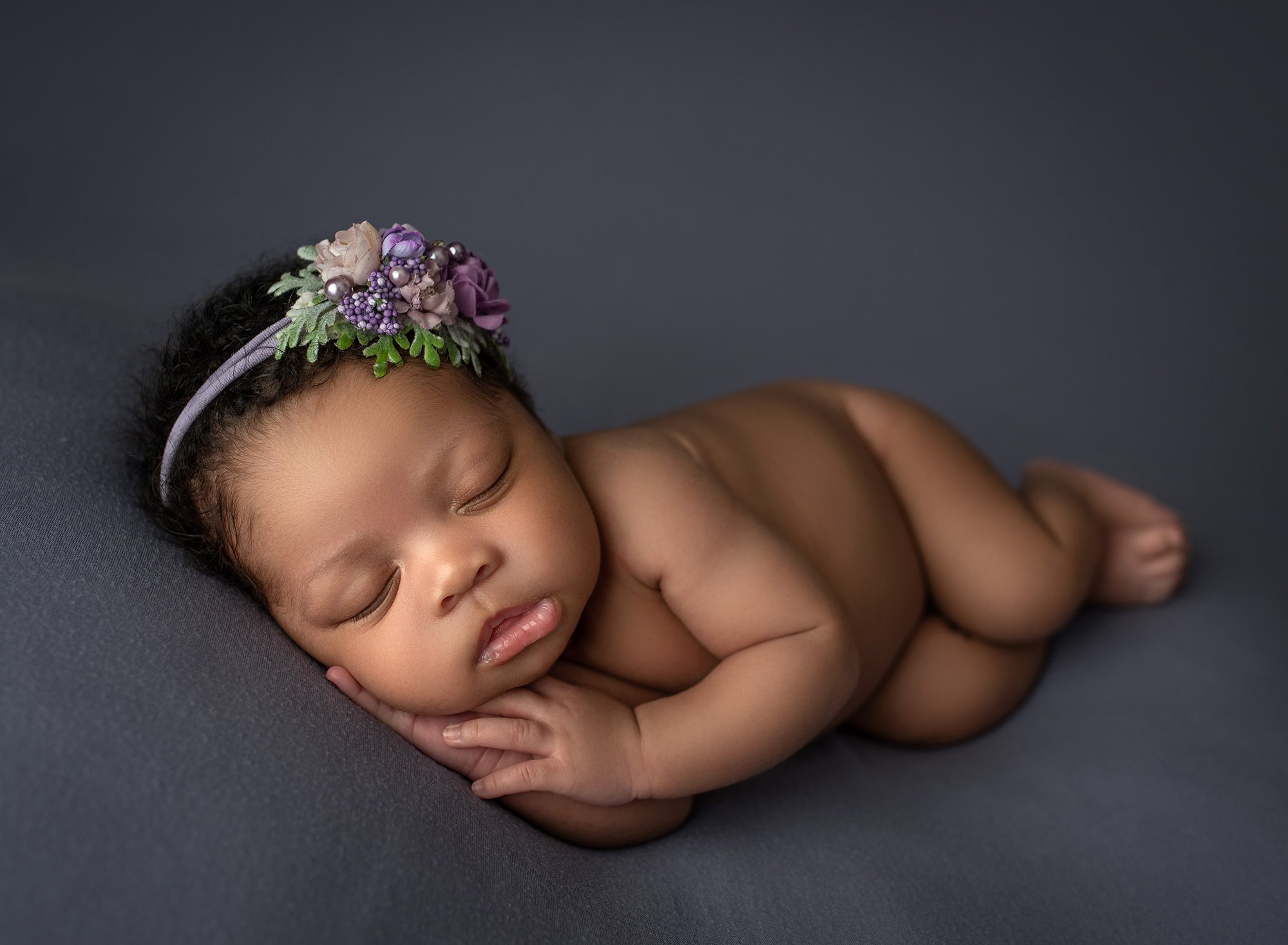 Best age for newborn photos naked baby girl laying on her side with a purple floral headband and her hands under her cheek