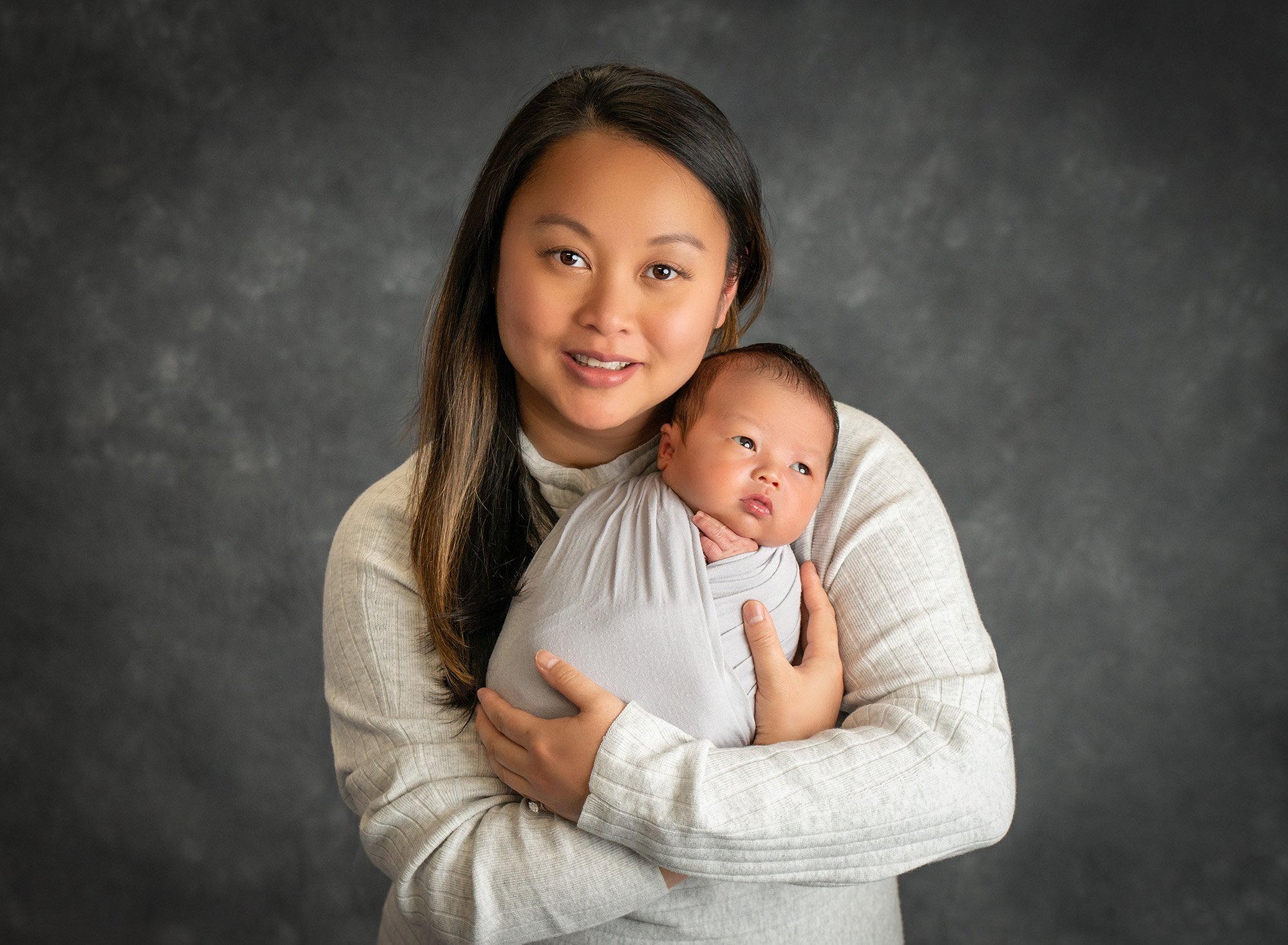 mom wearing cream sweater holding newborn son swaddled in gray