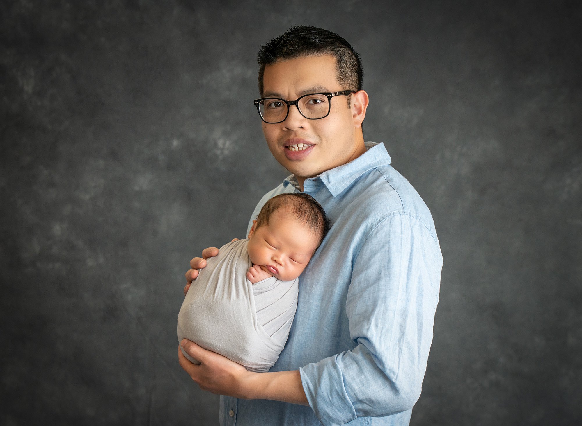 dad in blue button up holding newborn son swaddled in gray