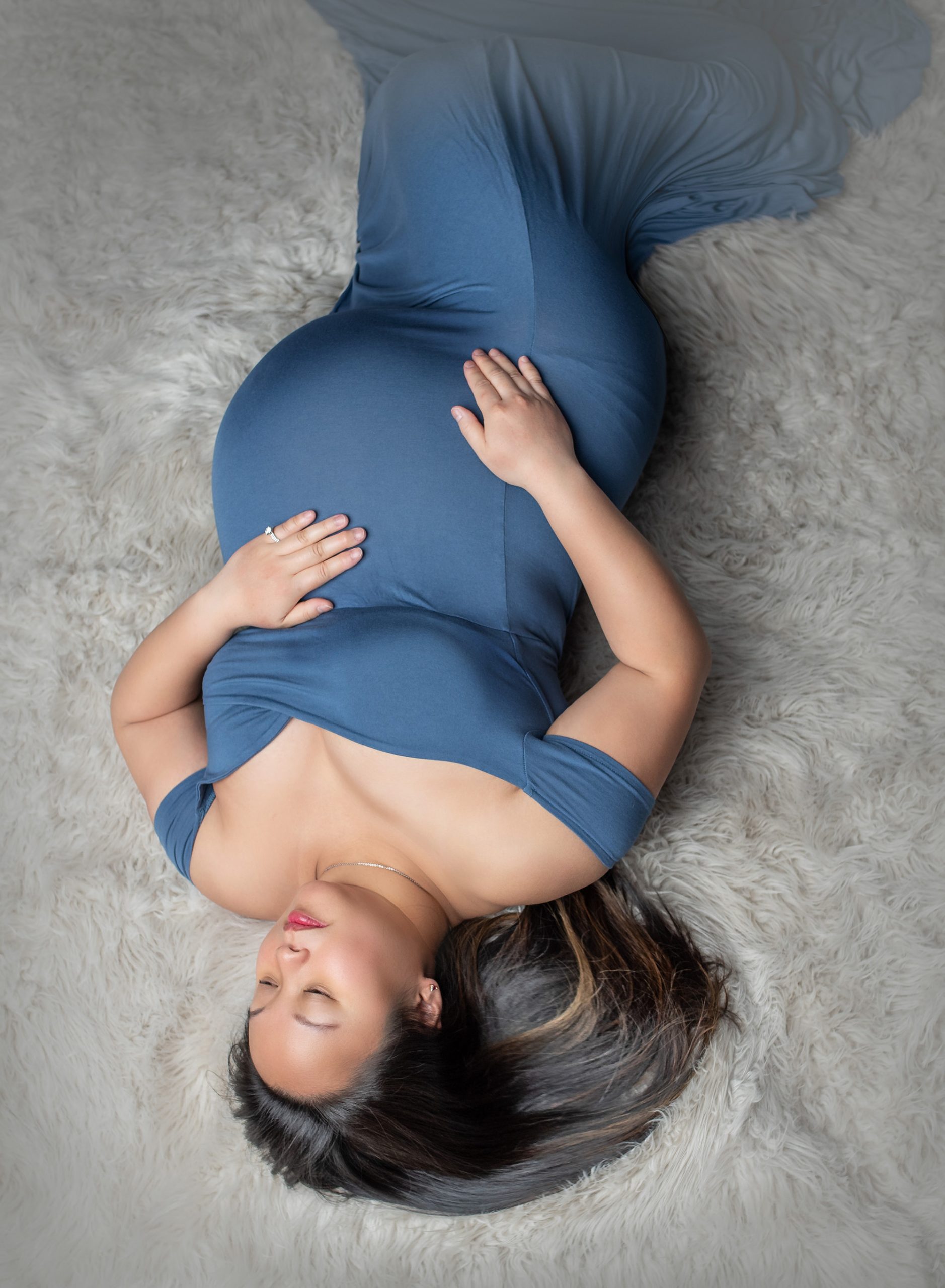 pregnant mother in blue maternity dress laying on her back on top of gray fluffy blanket
