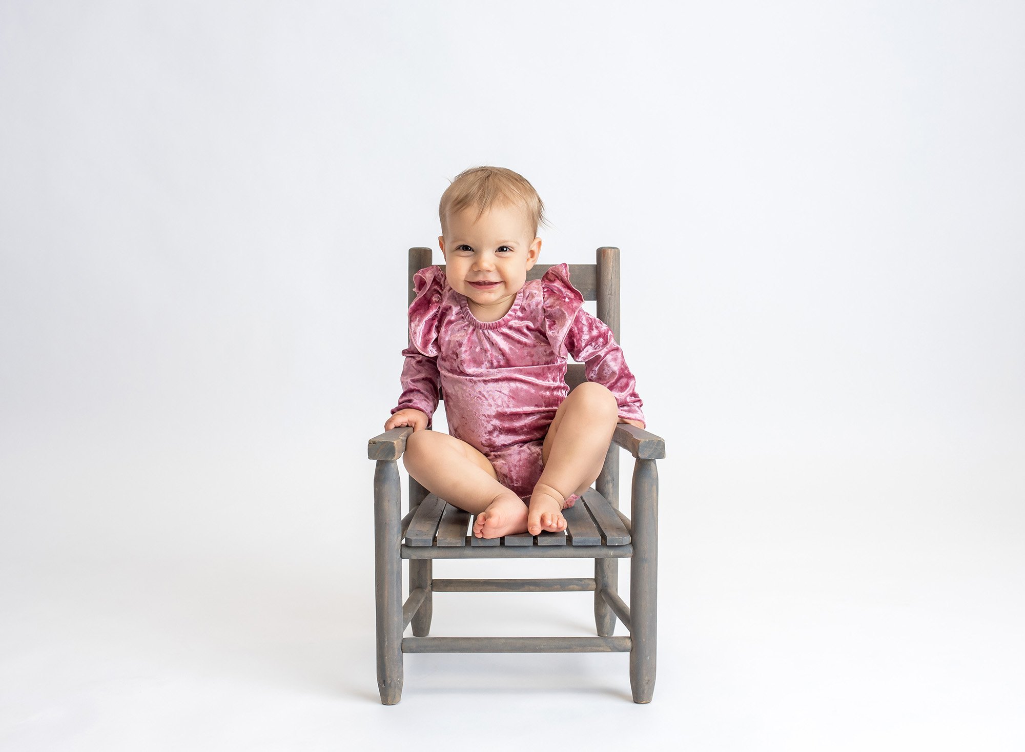 blonde one year old girl wearing pink velvet body suit sitting on rustic chair with white backdrop