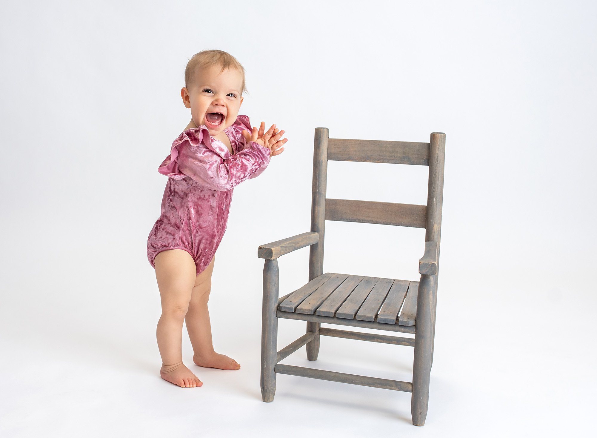 1 year baby photoshoot one year old girl laughing and clapping wearing a pink velvet ruffled body suit with rustic chair