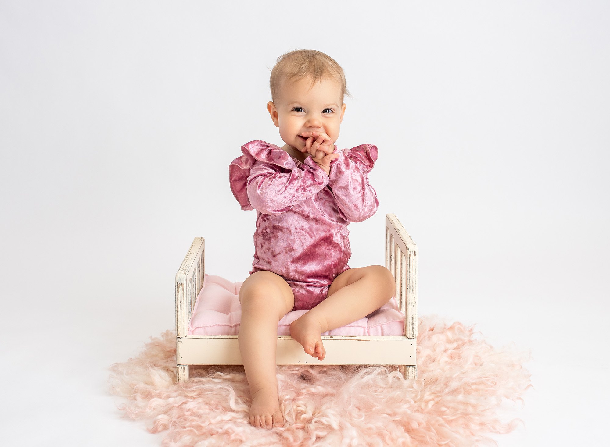 one year old girl wearing pink velvet ruffled bodysuit sitting on top of miniature white shabby bed with pink cushion