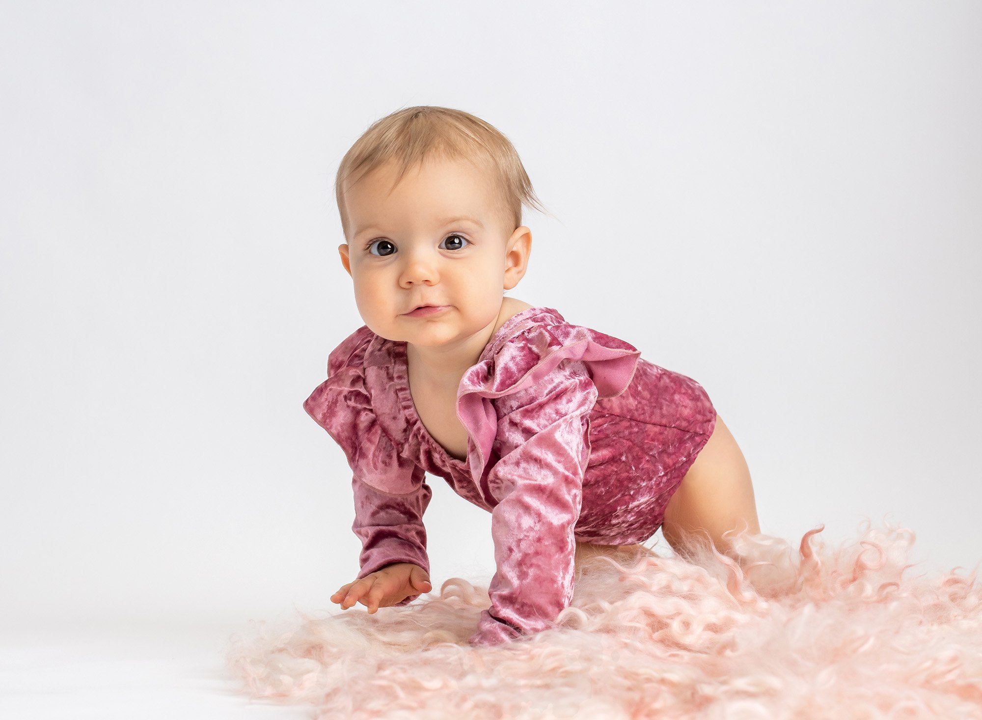 brown eyed one year old girl wearing pink velvet ruffled bodysuit on top of pink fuzzy blanket