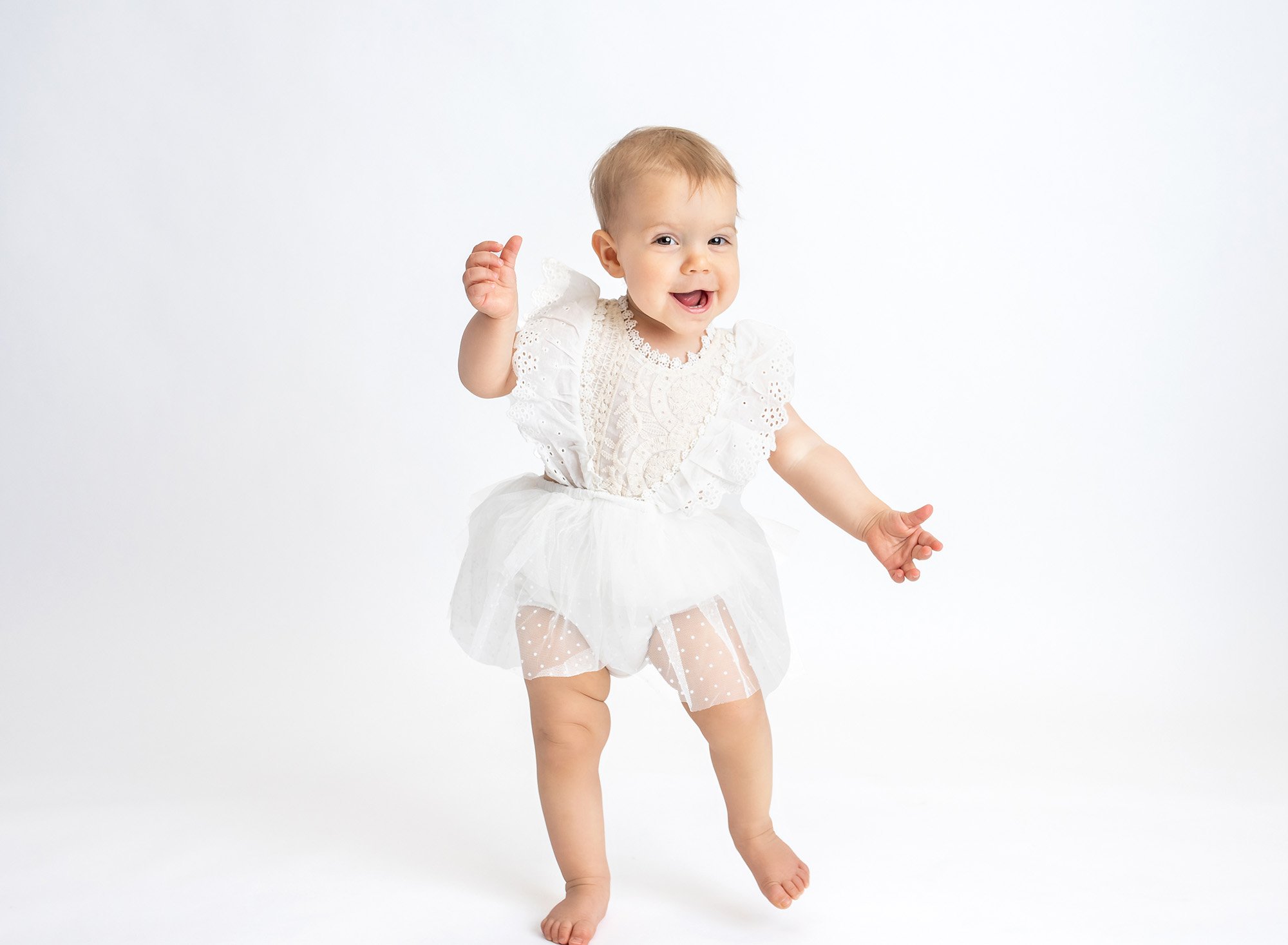 brown eyed one year old girl wearing white lace dress smiling and walking with white backdrop