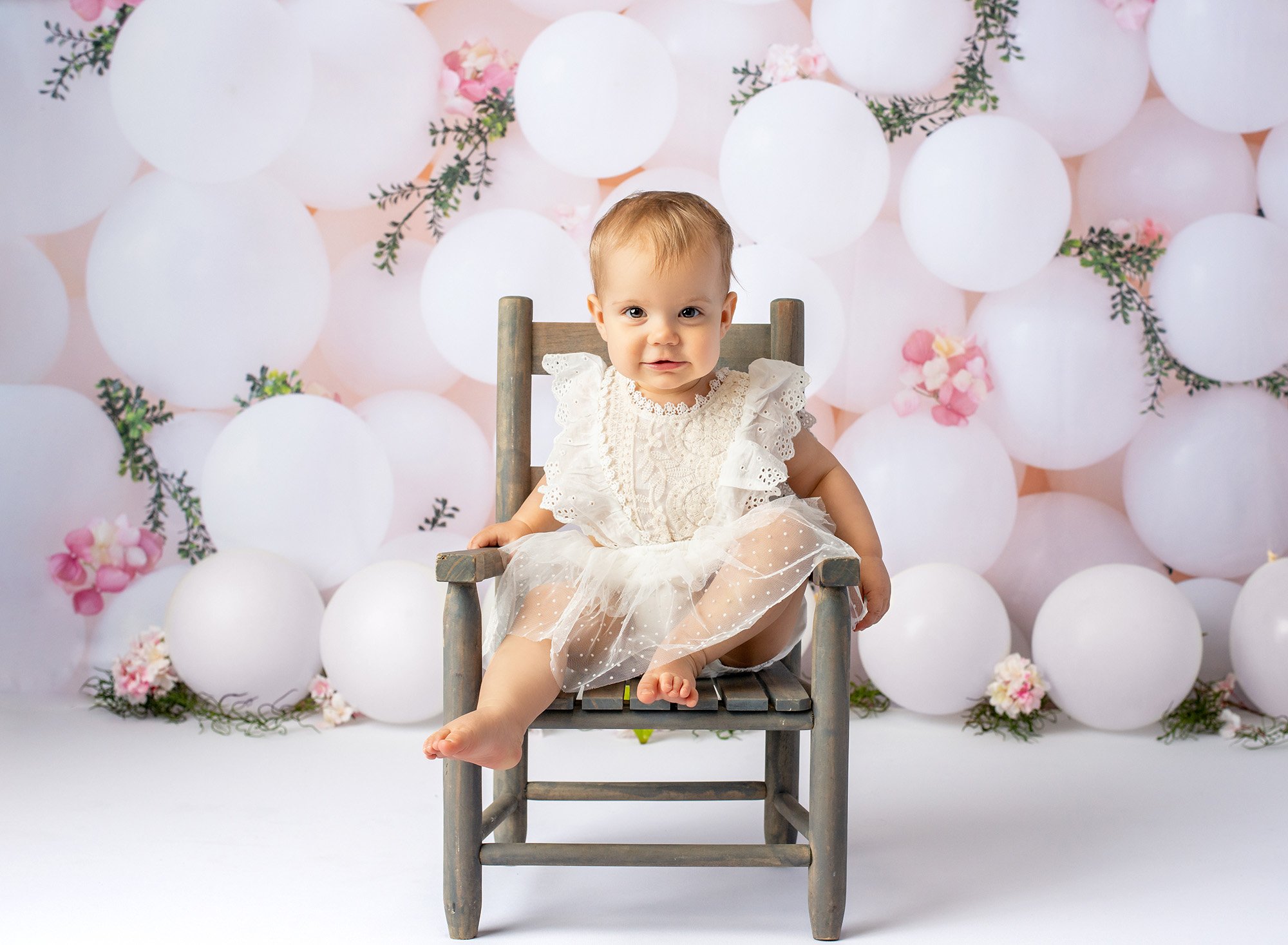 1 year baby photoshoot brown eyed one year old girl wearing white lace dress sitting on rustic chair with white balloons in background
